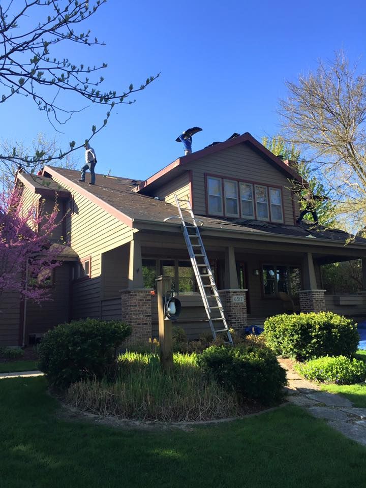 Two workers repair the roof of a two-story brown house with a prominent ladder resting against the front gable.