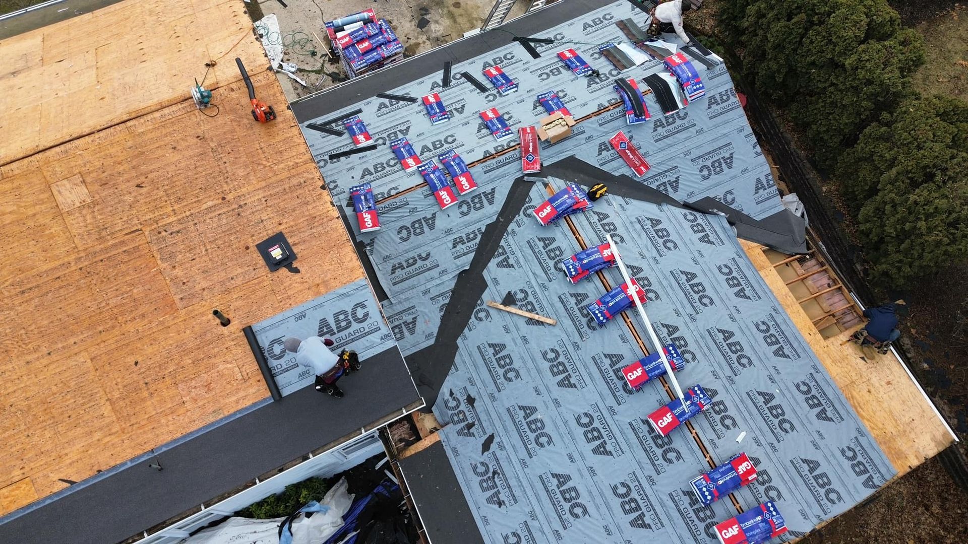 An aerial view of a roof under construction, showing workers installing gray ABC Supply underlayment over plywood decking.