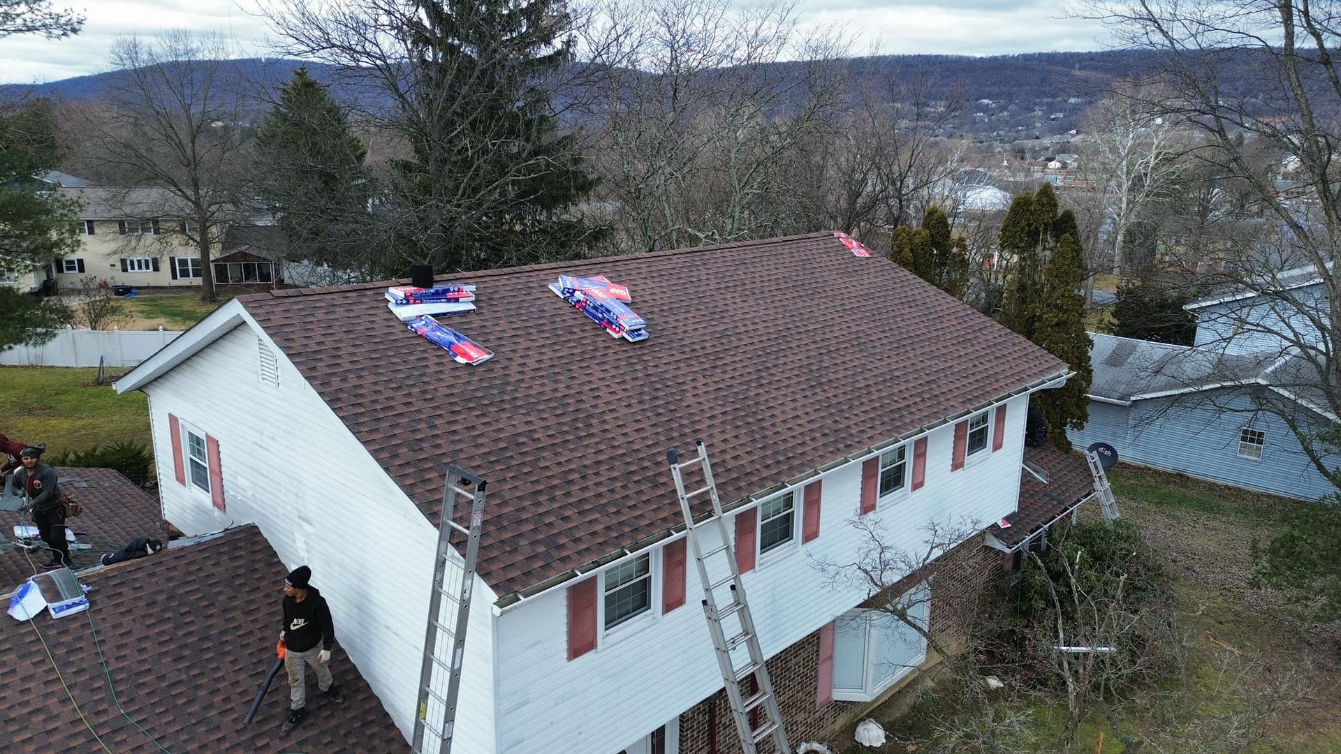 Workers install brown shingles on the roof of a two-story white house with red shutters.