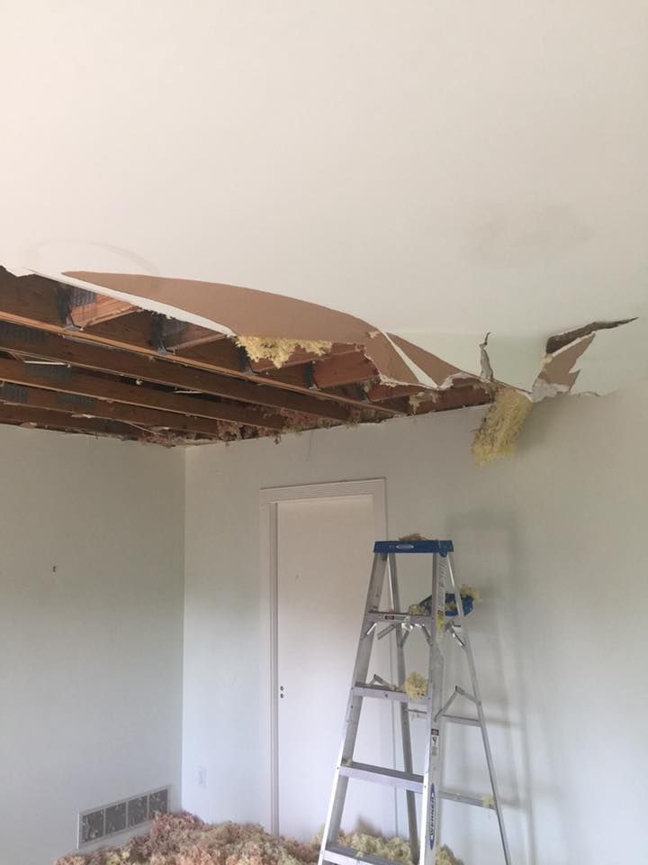 A room with a large section of ceiling drywall collapsed, exposing the wooden joists and insulation above a step ladder.
