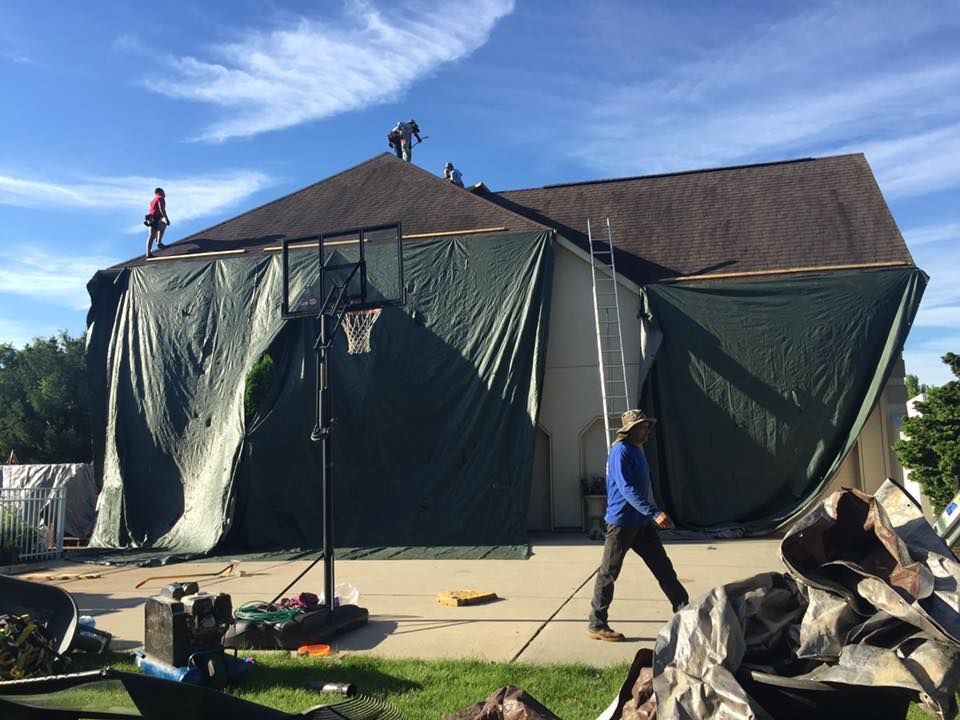 Workers on a house roof covered with green tarps during a renovation project, with a basketball hoop in the foreground.