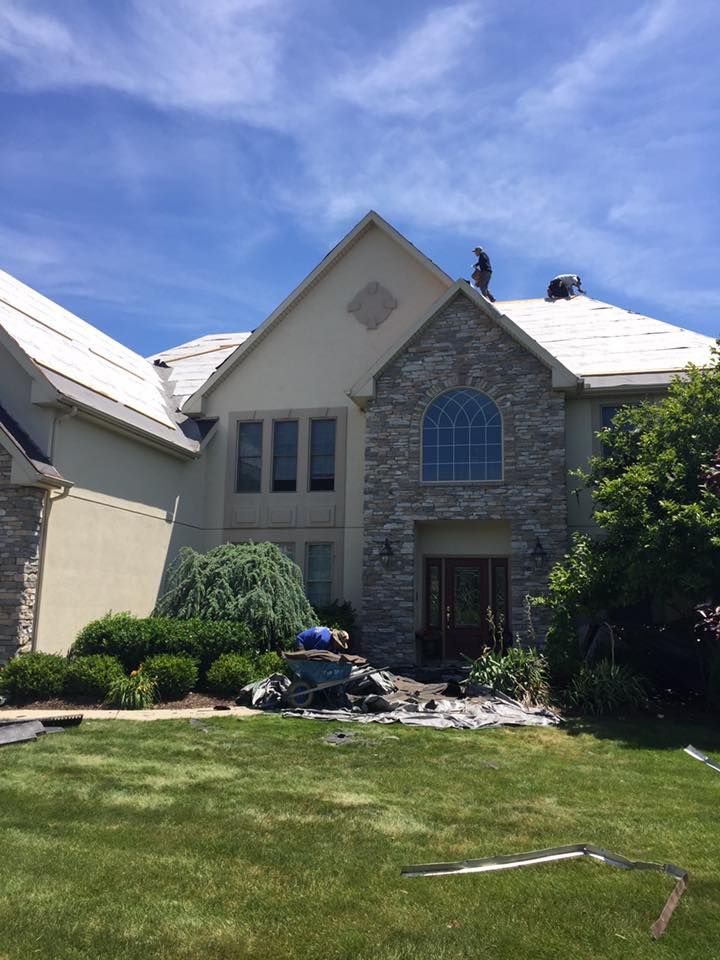 Roofers work on the steep roof of a two-story stone and stucco house under a bright blue sky.