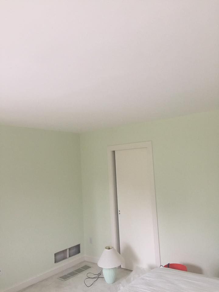 A corner of a room with pale green walls, white trim, a closed door, a floor vent, and a small table lamp on the floor.