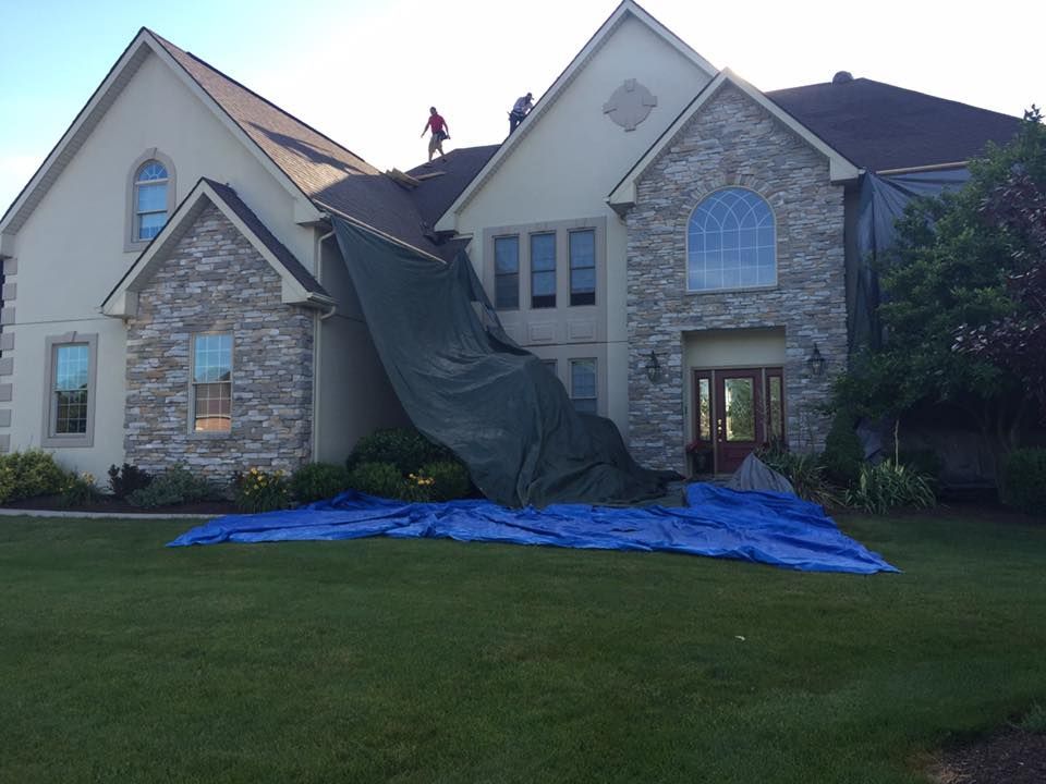 Workers on the roof of a large house with a protective tarp covering one side and a blue tarp spread on the lawn.