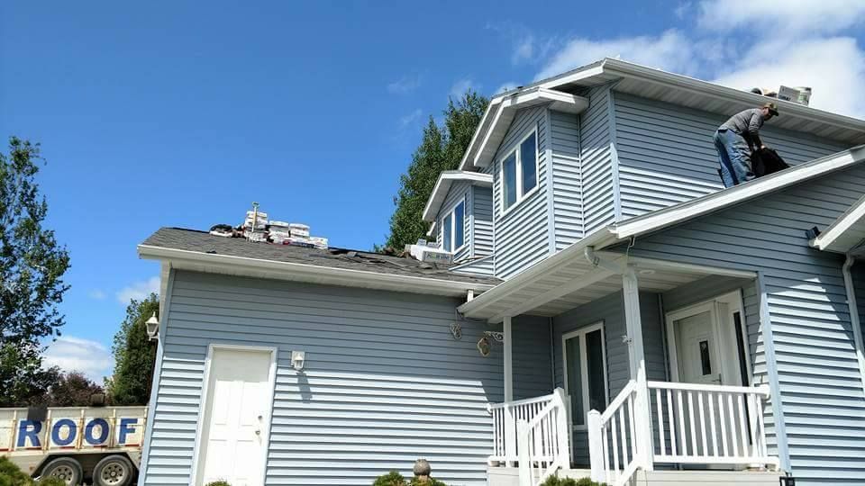 A worker installs a new roof on a light blue, two-story house under a bright blue sky, with a roof supply trailer nearby.