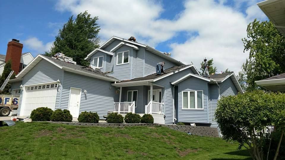 A two-story, light blue house with a white garage and porch sits on a grassy, sloped lot under a cloudy blue sky.