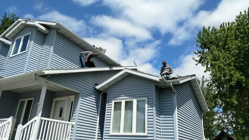 Two workers on the roof of a two-story blue house under a cloudy sky.
