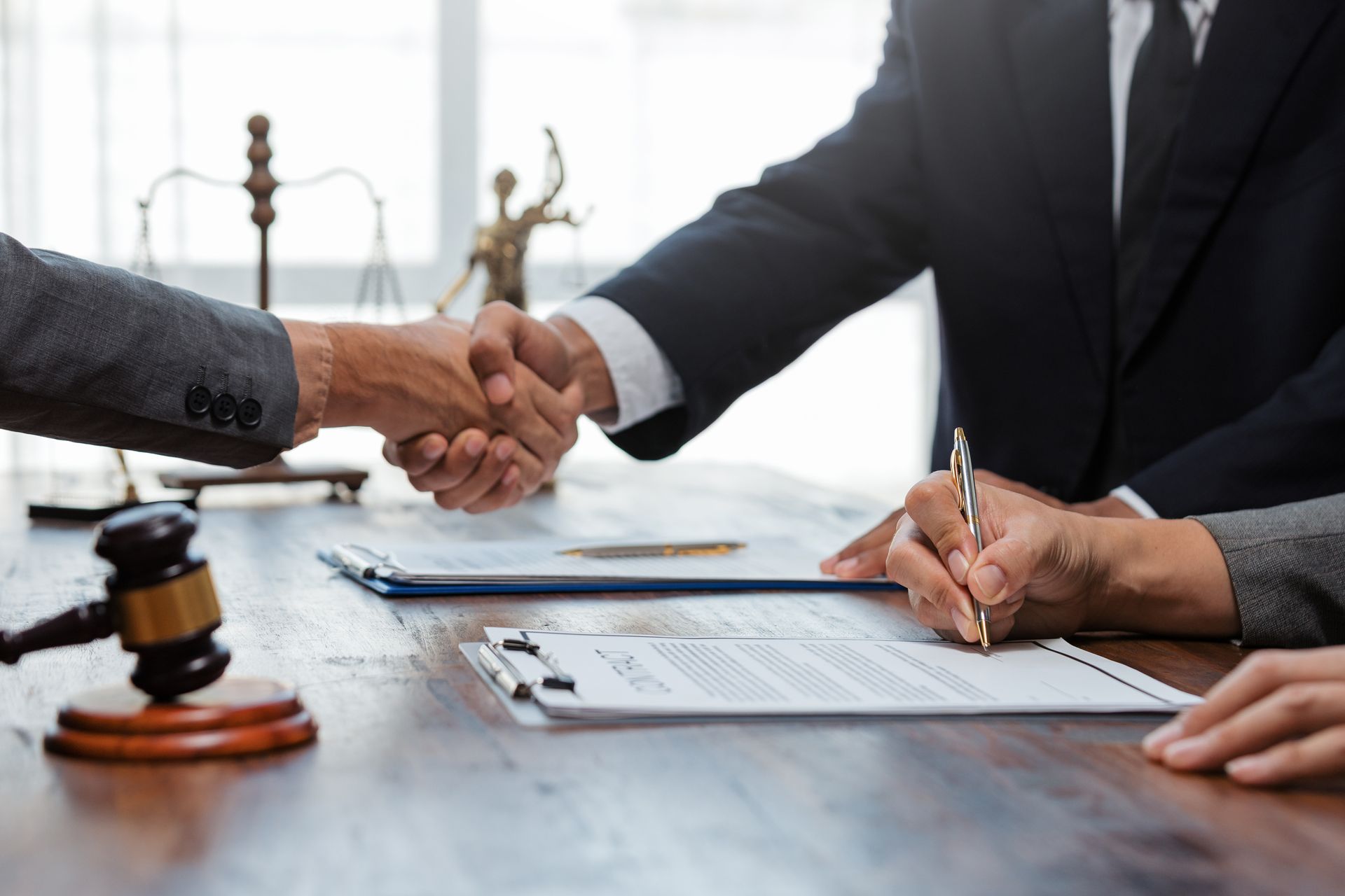 A lawyer and a client shaking hands across a desk, with another lawyer signing a document. A lawyer and a client shaking hands across a desk, with another lawyer signing a document.