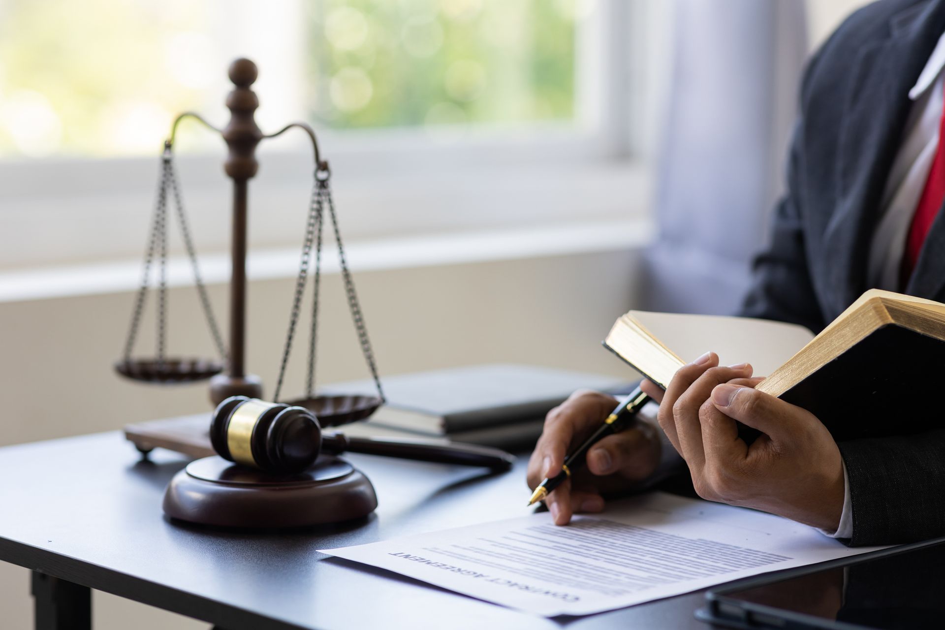 A lawyer in a suit reviews documents, holding a pen and book, with scales of justice and a gavel on the desk.