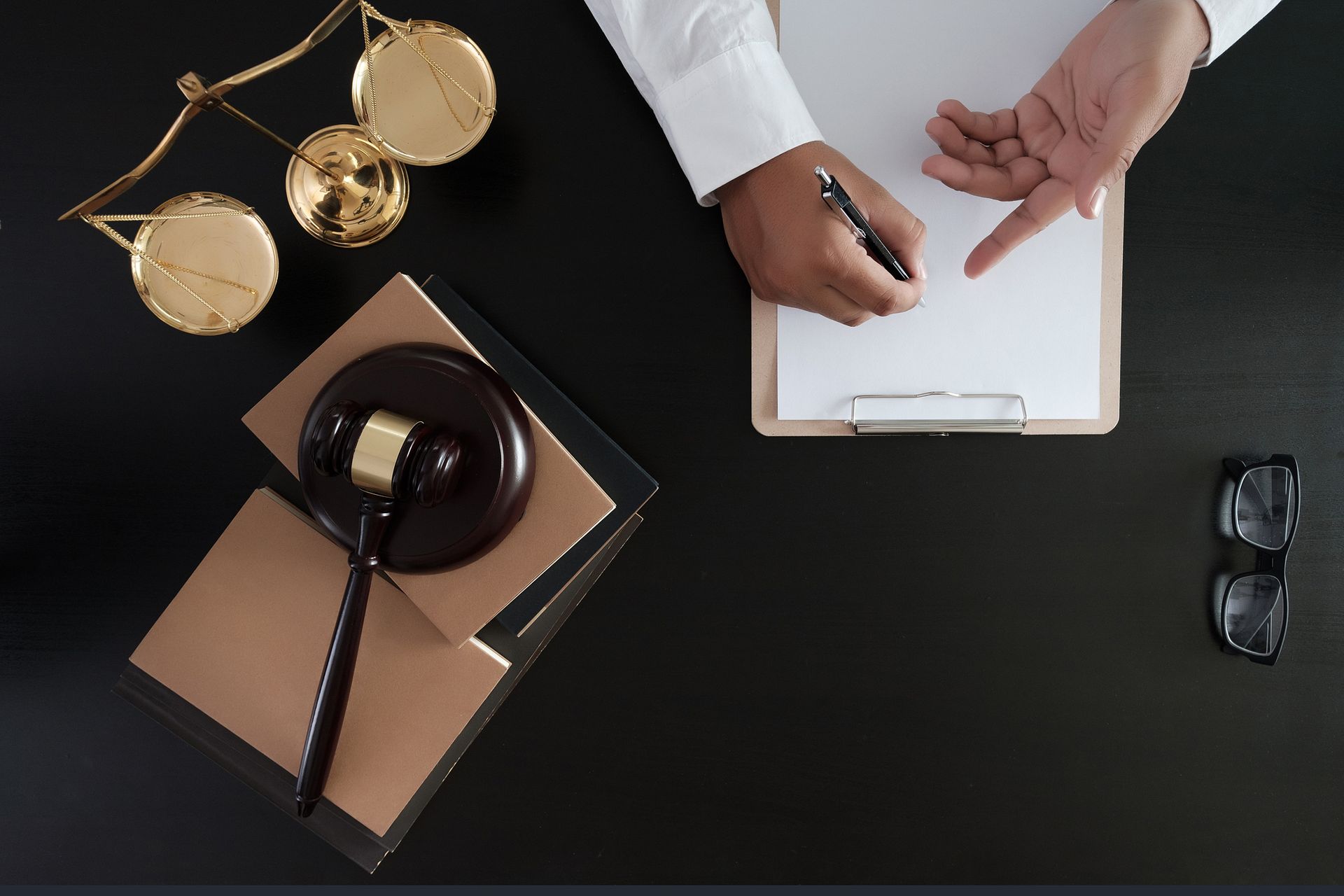 Criminal solicitor discussing legal case with gavel, scales, glasses, and clipboard on black desk. 