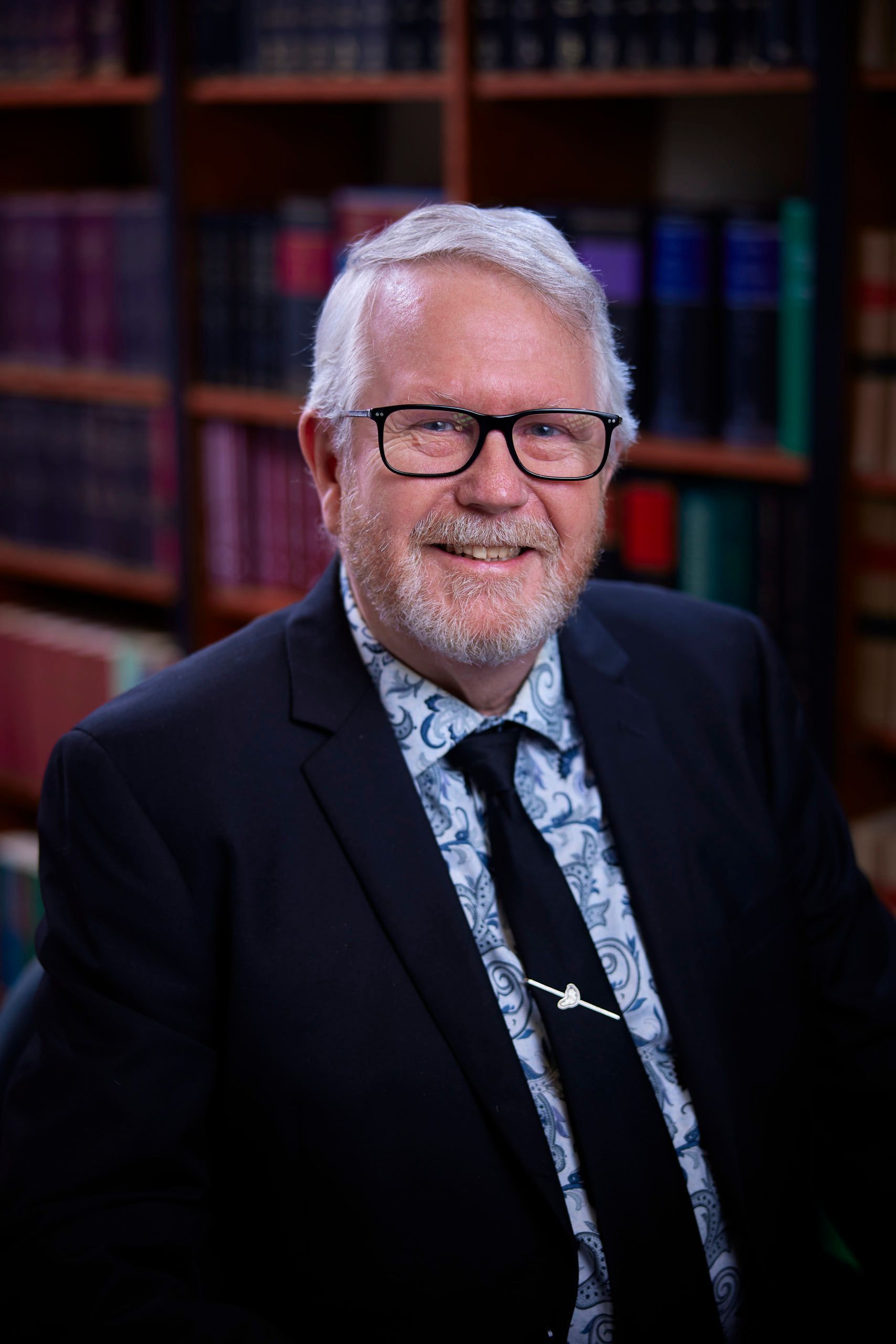 A man in a suit and tie is smiling in front of a bookshelf.