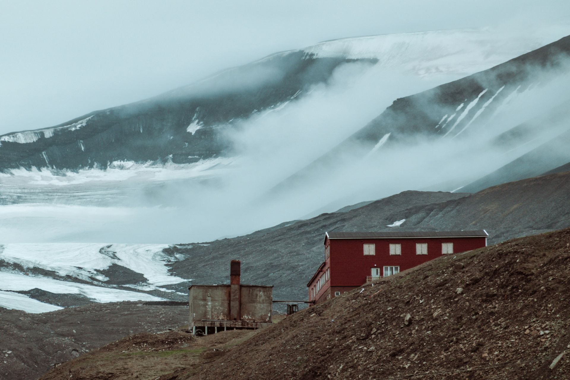 A red house is sitting on top of a hill in front of a snowy mountain.