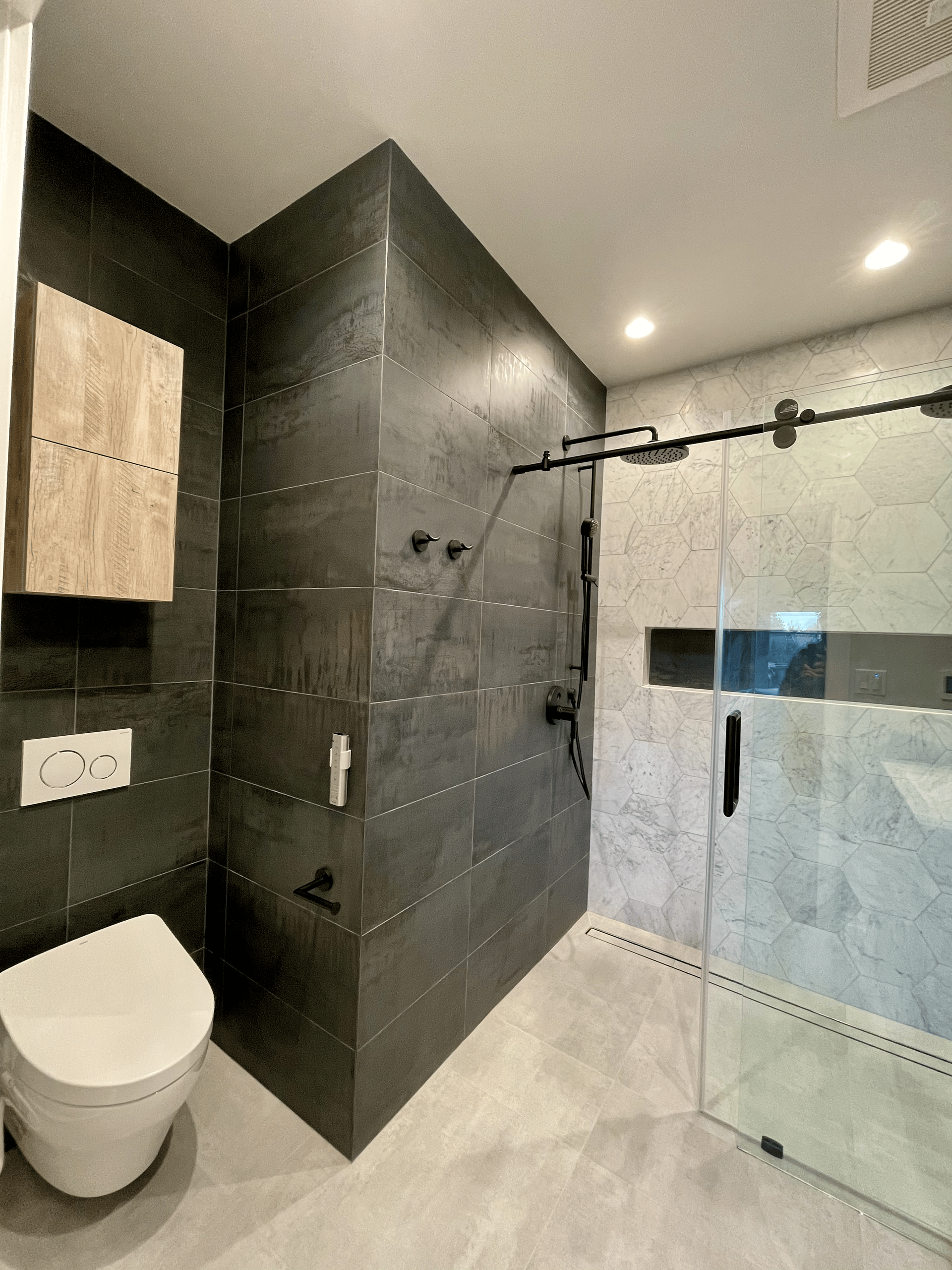 Modern bathroom with dark gray tiled shower and light gray floor. Toilet and light wood cabinet on the left.