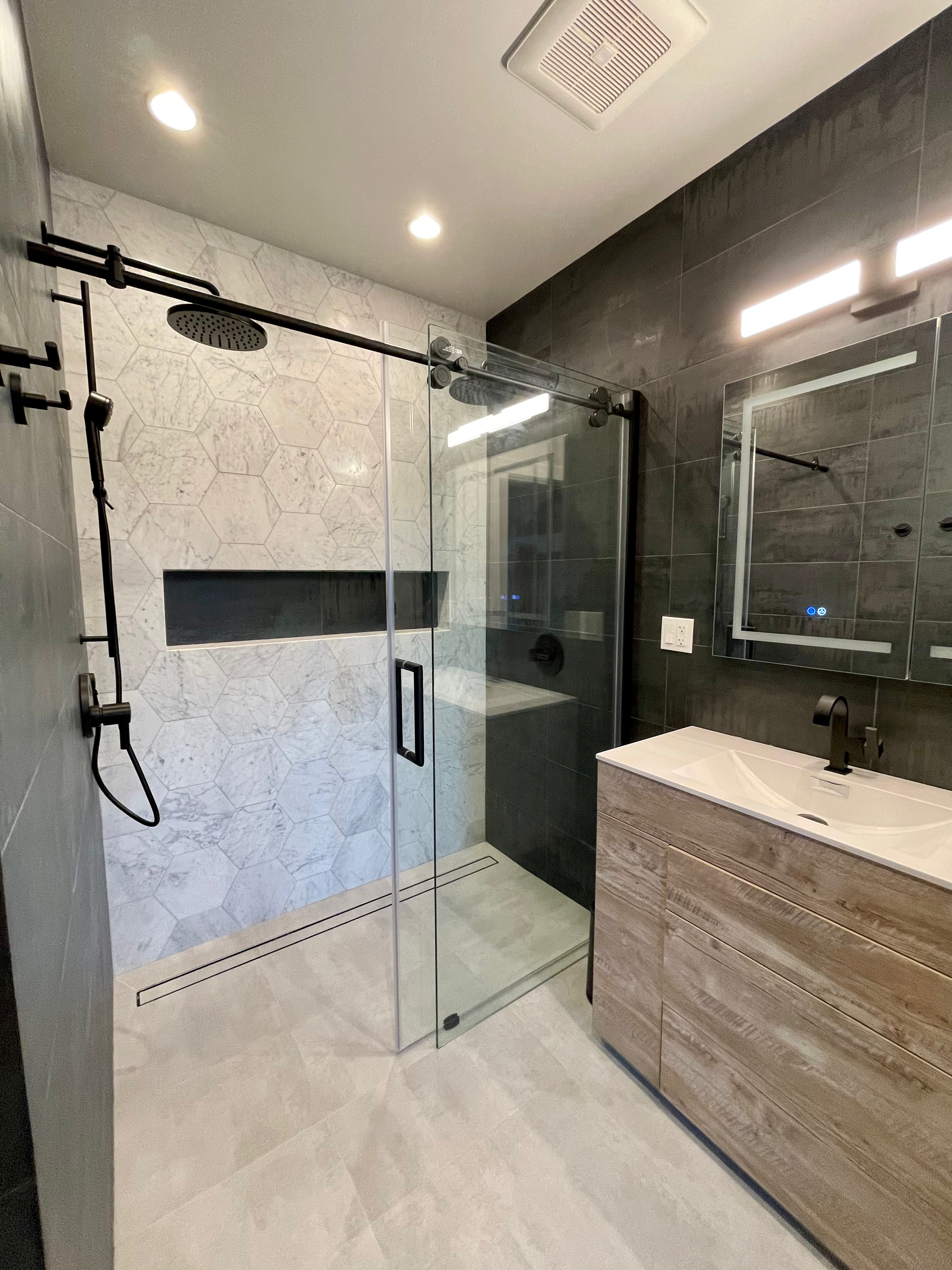 Modern bathroom with glass shower, gray tile, light wood vanity, and black fixtures.