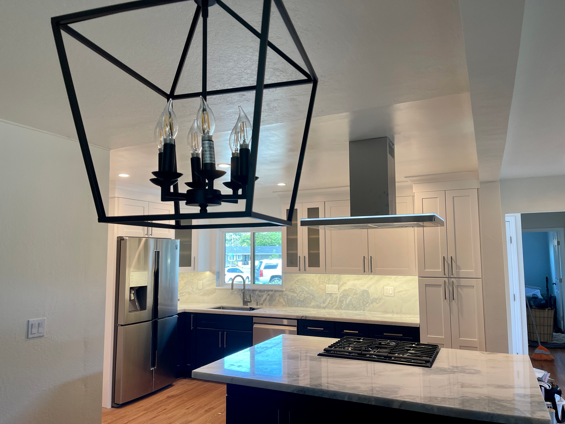 Black geometric chandelier hangs above kitchen island with stove. Stainless steel appliances and white cabinets.