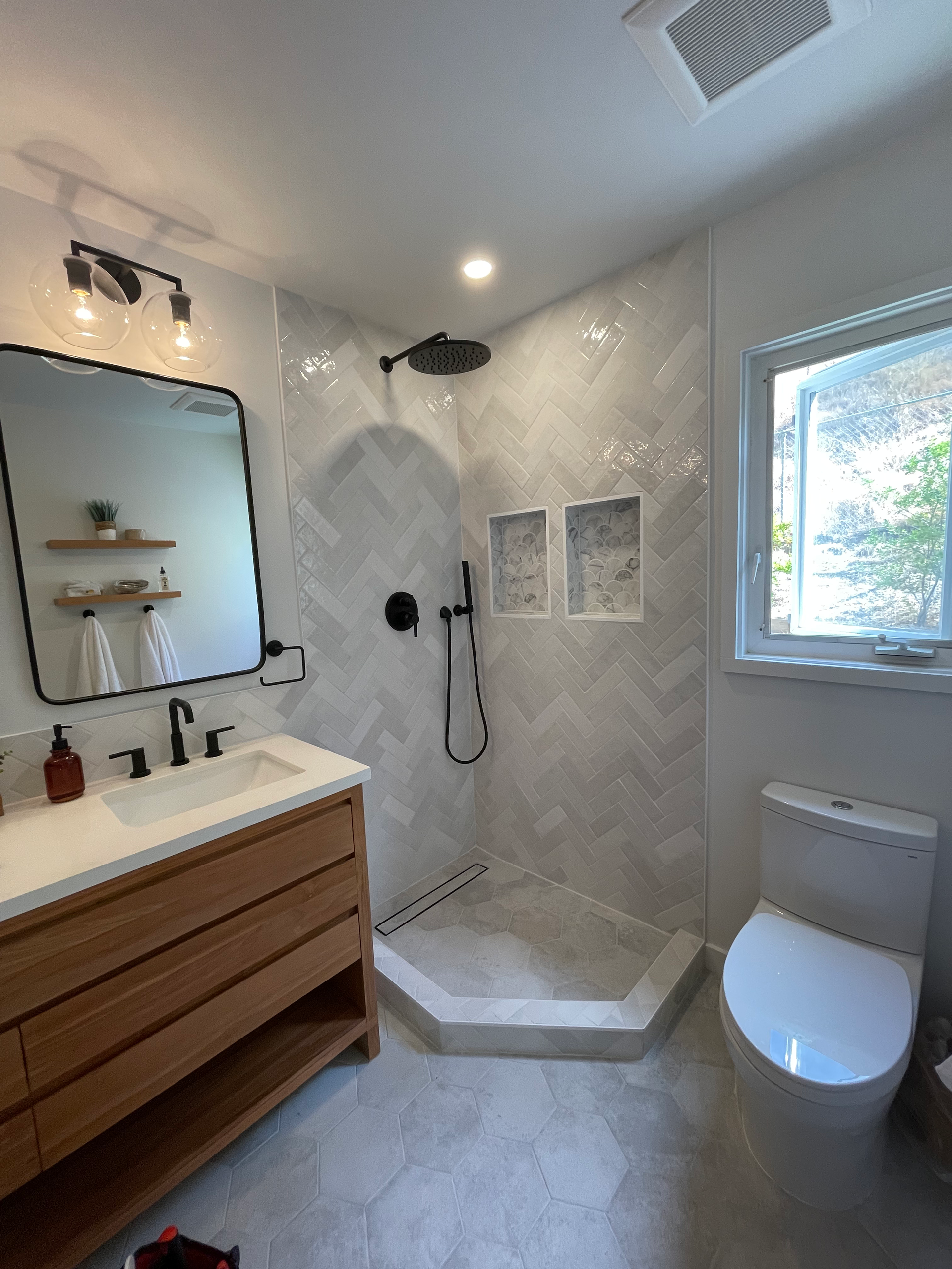 Modern bathroom with a wooden vanity, tiled shower, and toilet next to a window.