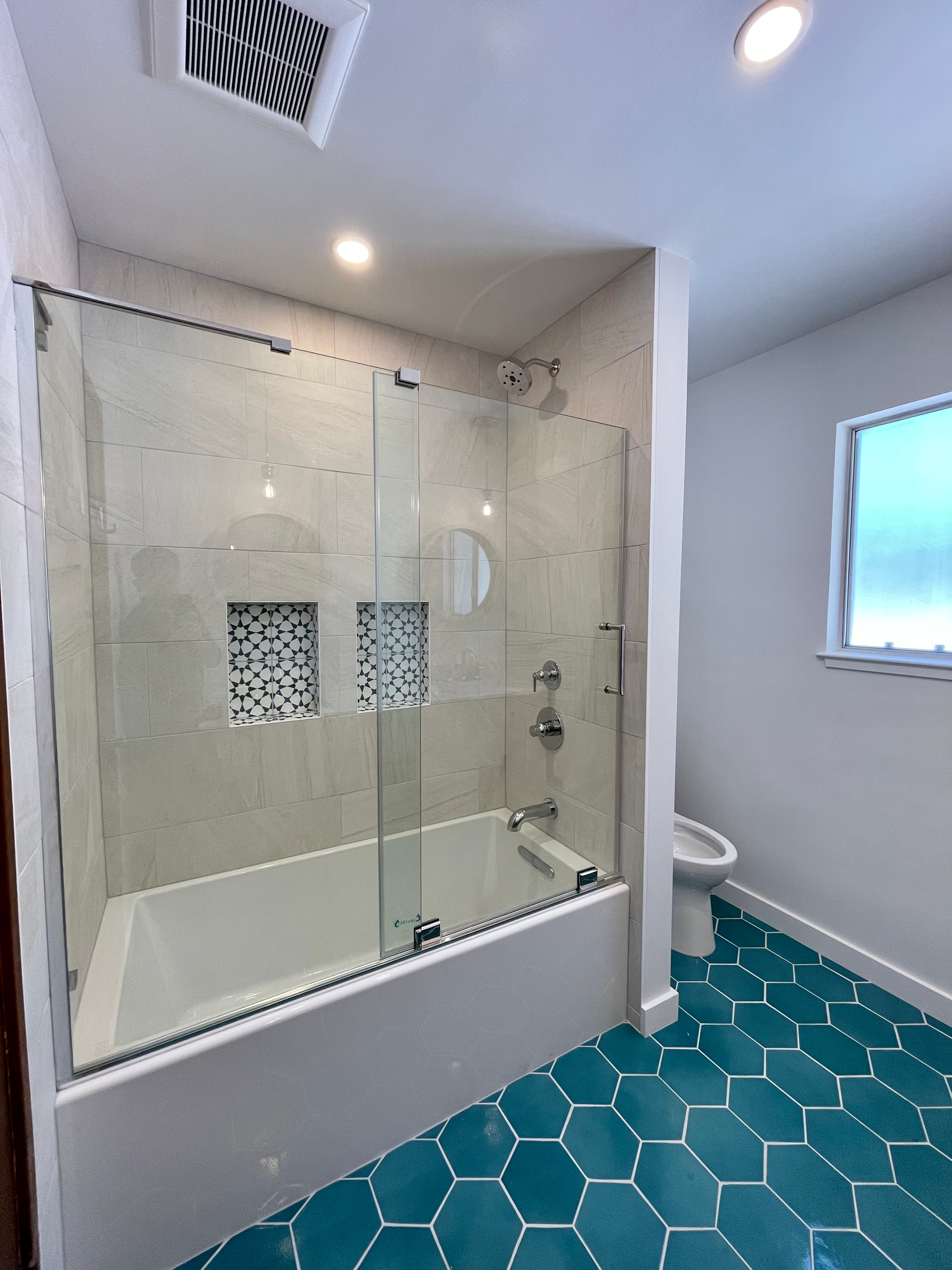 Bathroom with teal hexagon tile floor, white tub, glass shower doors, and light gray tile walls.