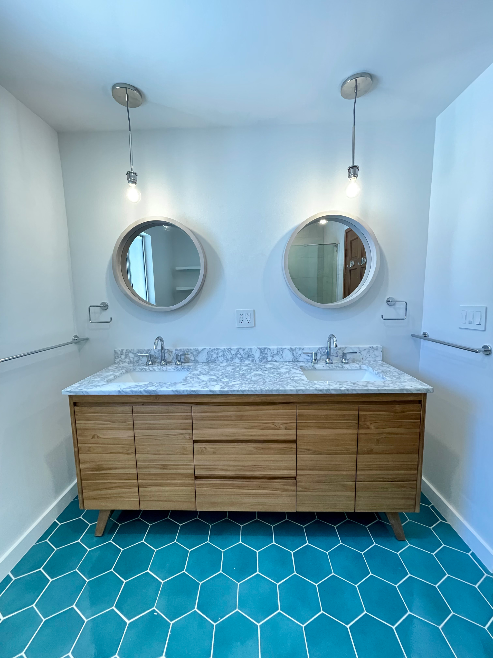 Bathroom with turquoise honeycomb tile floor, light wood vanity, round mirrors, and hanging lights.