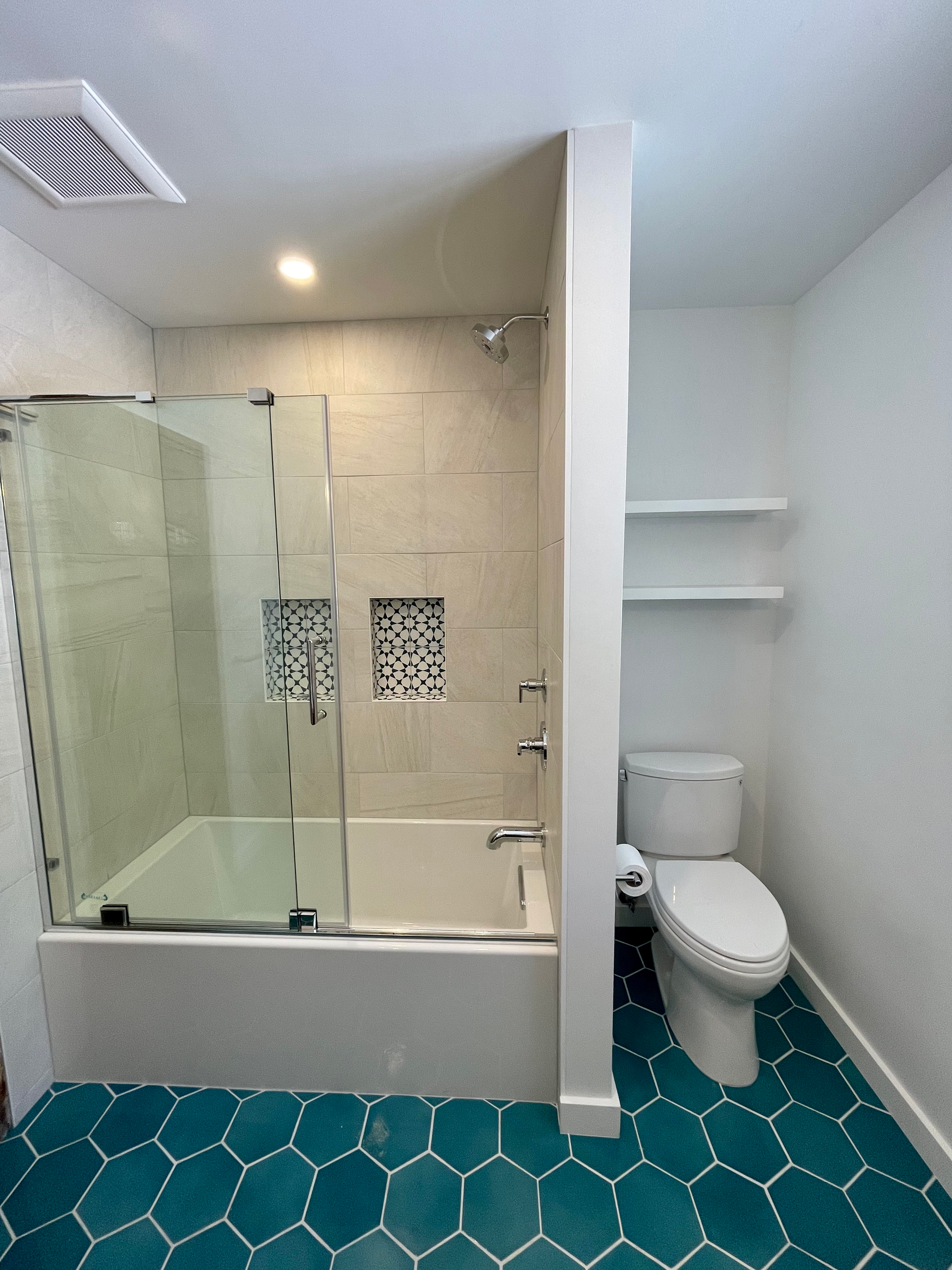 Bathroom with teal honeycomb tiles, white walls, shower over tub, and a toilet next to shelves.