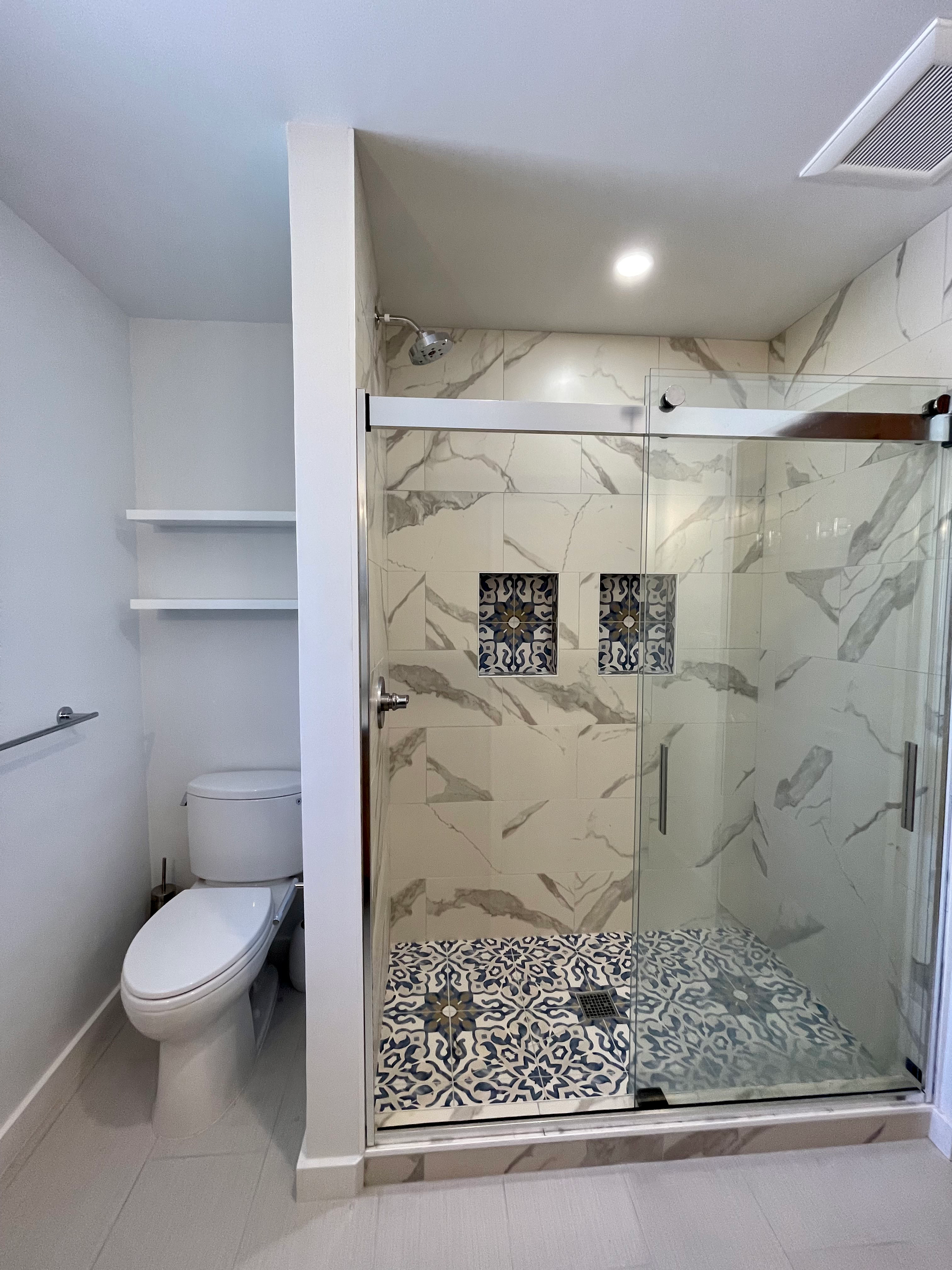 Modern bathroom with a toilet, shower, and shelves. The shower has decorative tile accents.