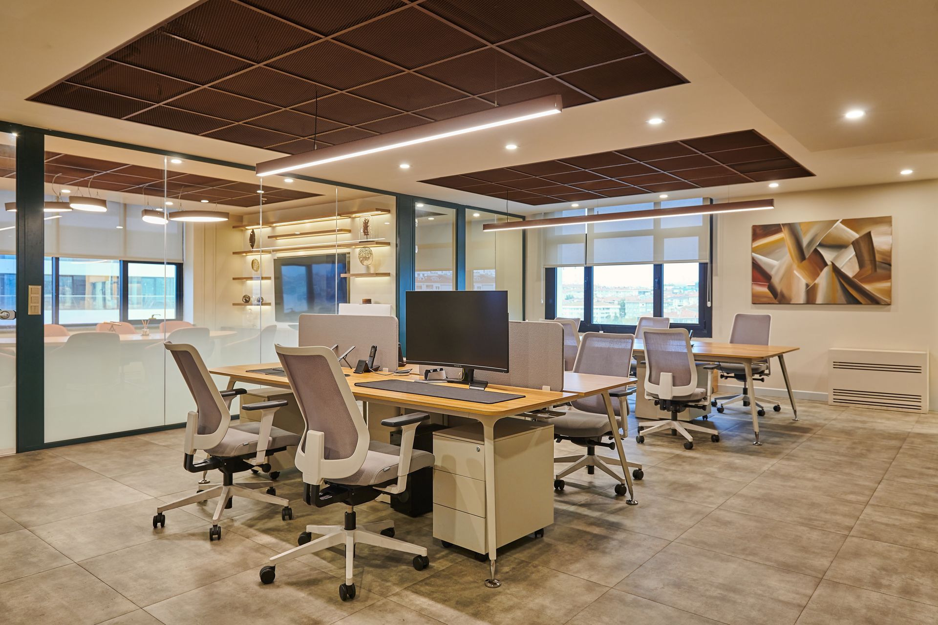 Modern office workspace with desks, swivel chairs, monitors, and large windows under recessed lighting