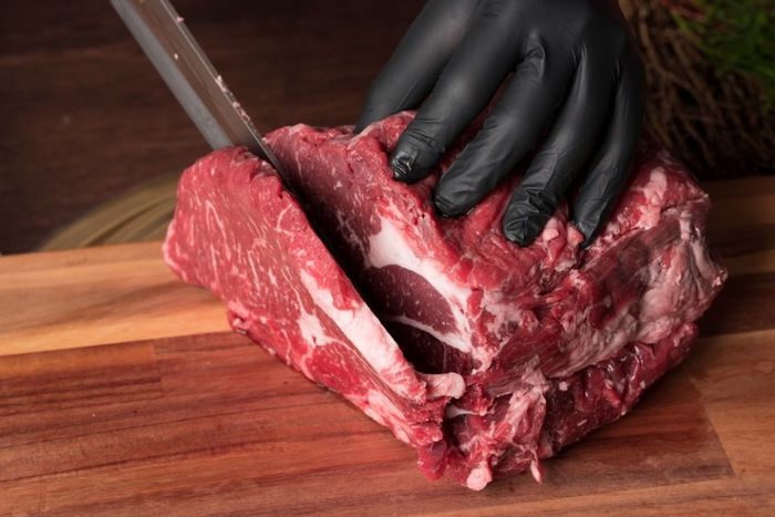 The butcher wearing black gloves cuts the meat with a knife on a wooden cutting board.