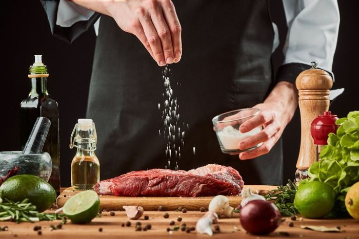 Man sprinkles salt over meat. Chef cooking a beef steak, seasoning with pepper, lime, olive oil, garlic and onion.