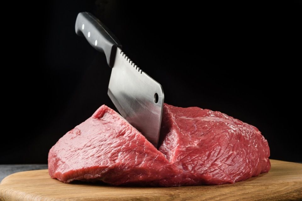 A piece of raw meat close-up with a chef's hatchet on a dark background.