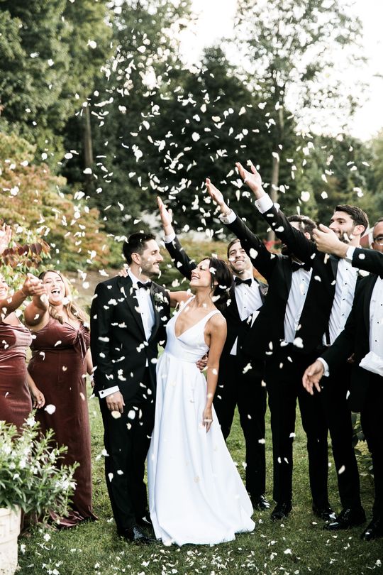 A bride and groom are surrounded by their wedding party throwing confetti in the air.