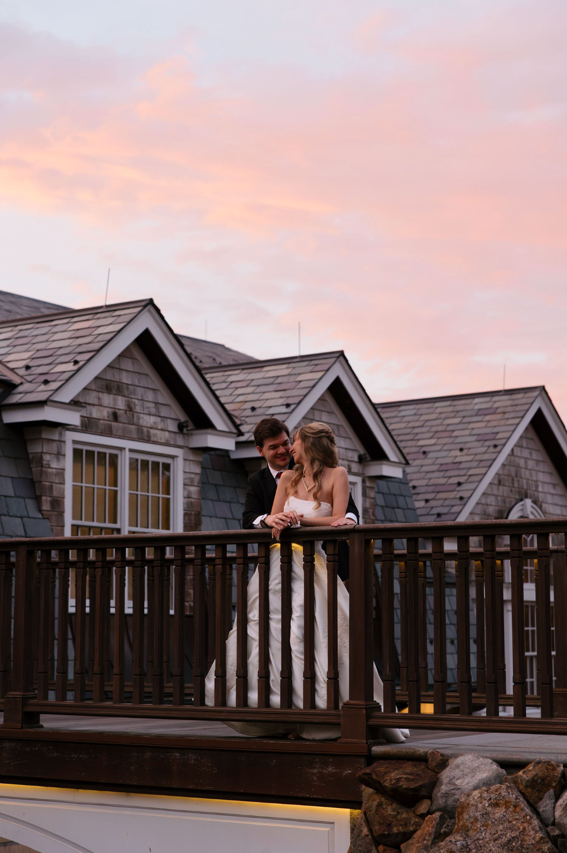 Bride and groom on a wooden deck at The Whittemore, looking at each other. Sunset colors in the sky and a building with gray shingles behind them.
