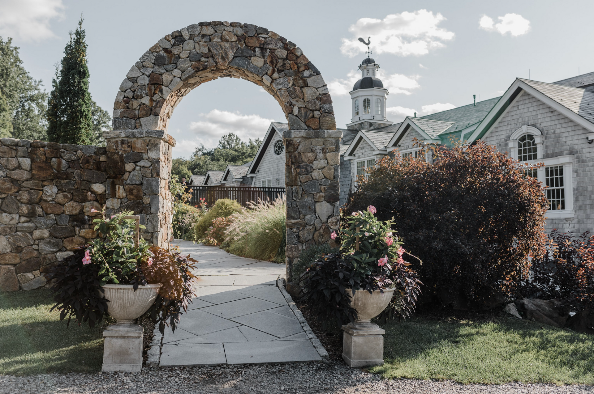 Stone archway with potted flowers leading to a path and buildings with a white-domed tower on a sunny day.