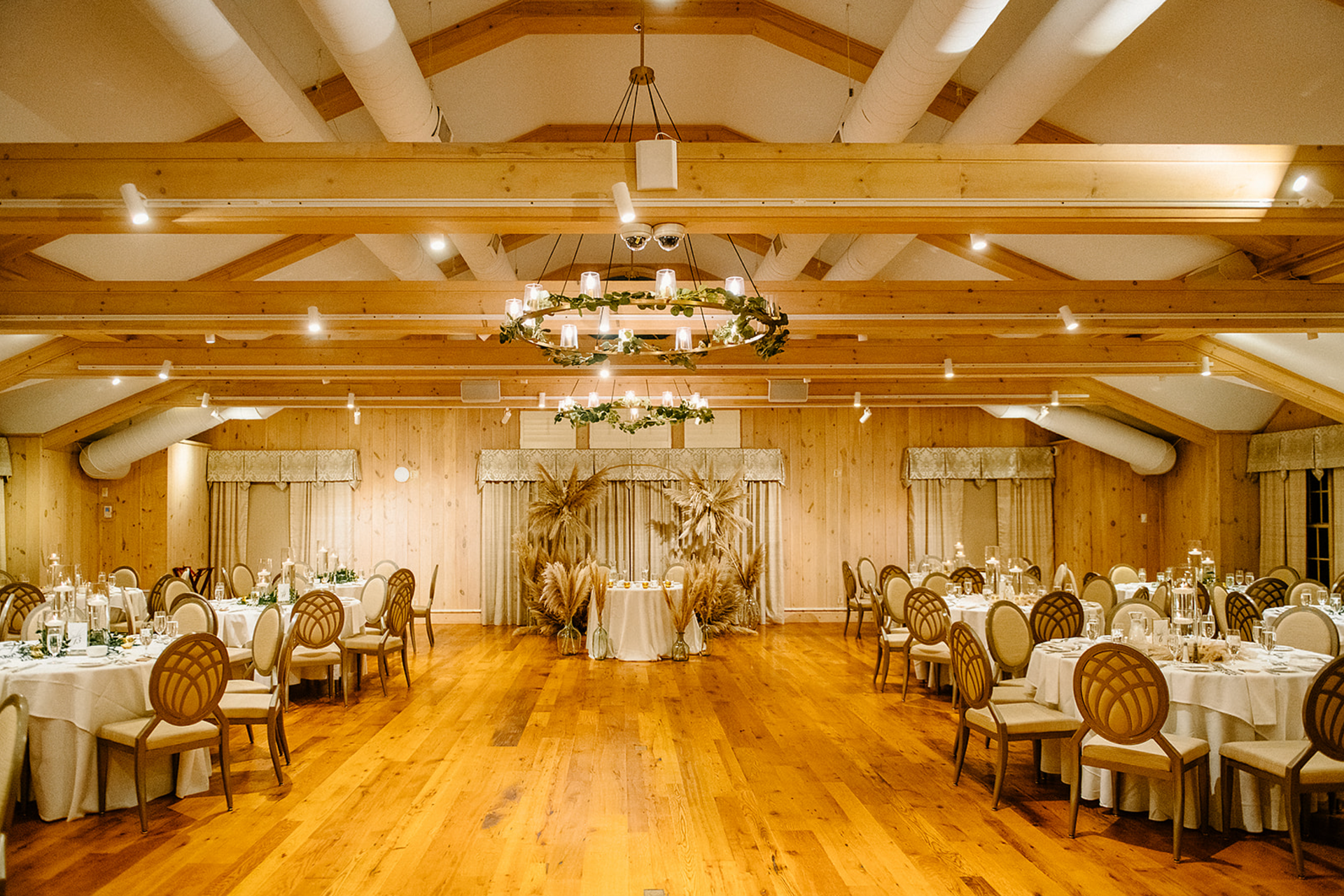Wedding reception venue with round tables, wooden floors, and a floral archway. The room has light wood beams and a chandelier.