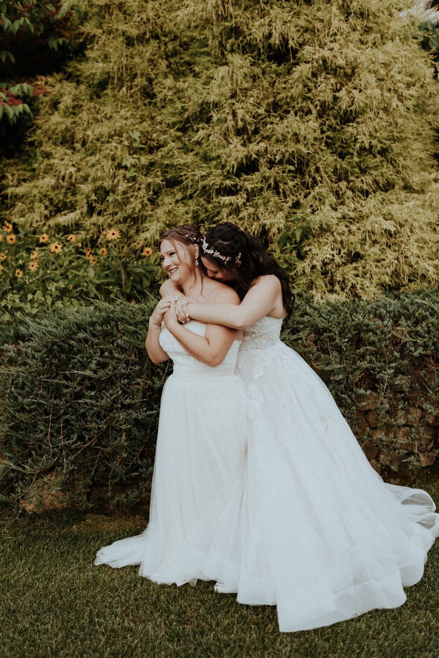 Two women in wedding dresses are hugging each other in front of a bush.
