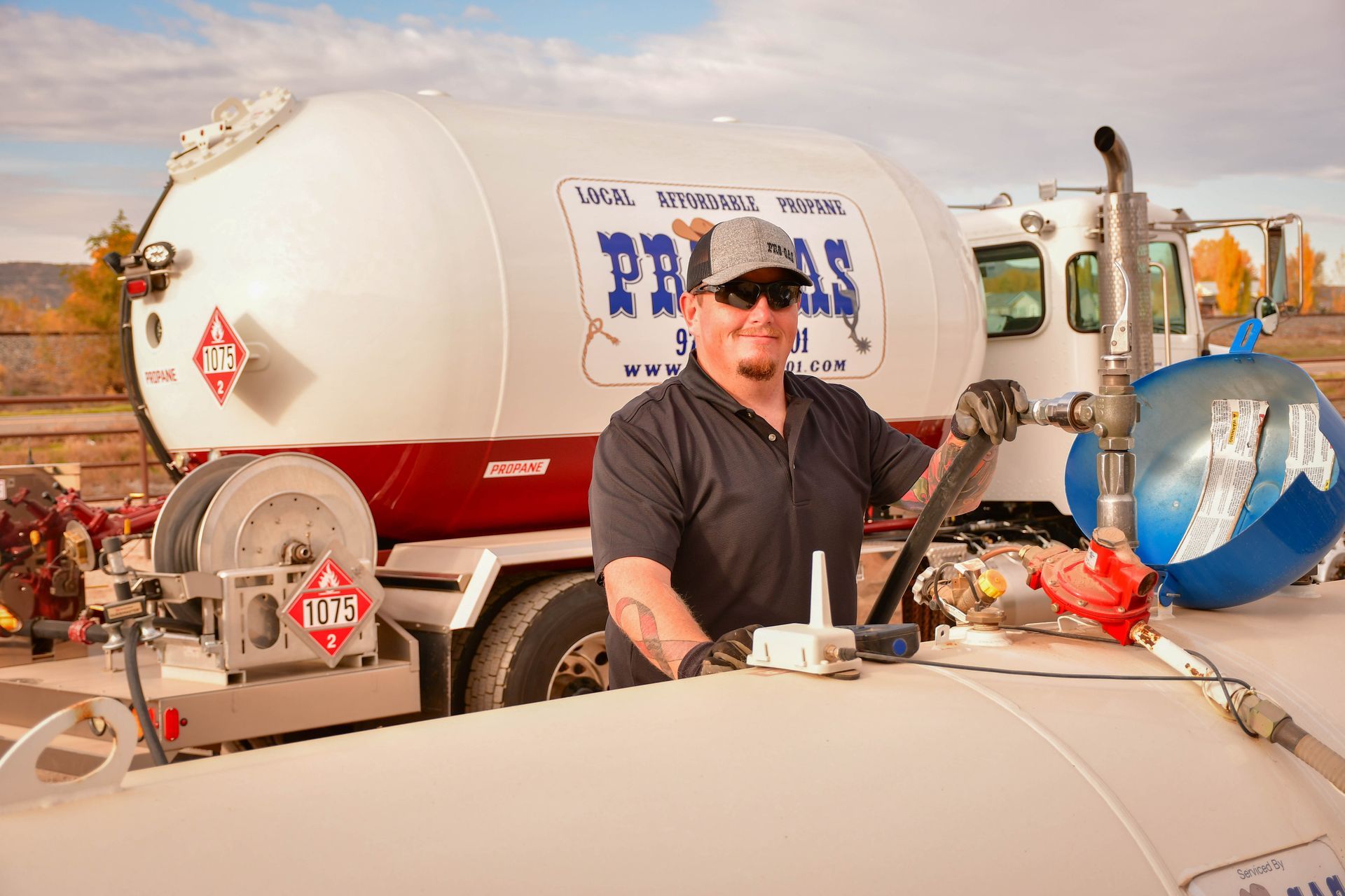 Man operating propane tank on truck, wearing gloves and sunglasses. White truck with red accents. Outdoors.