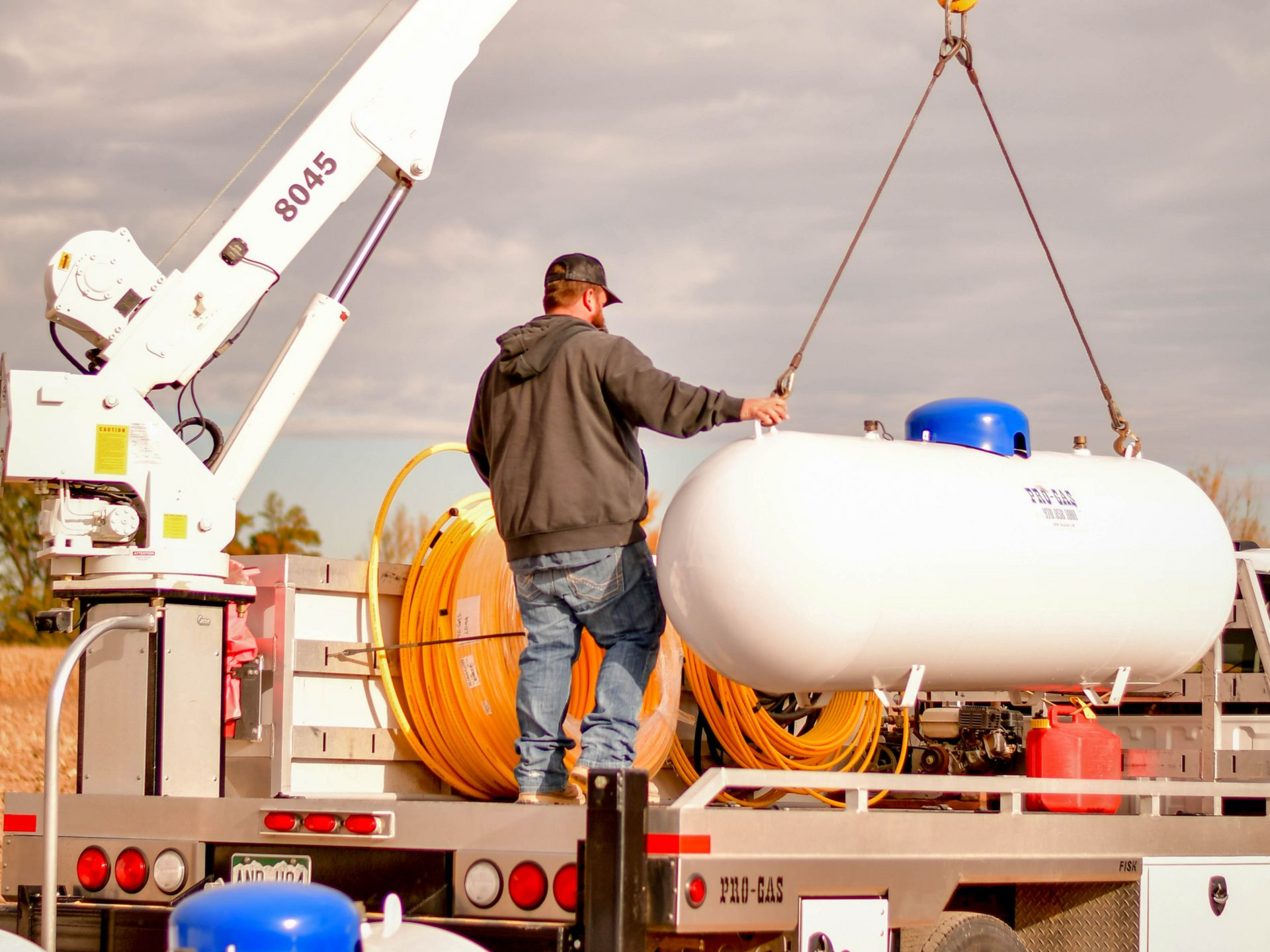 Man guiding propane tank being lifted by crane on truck bed.
