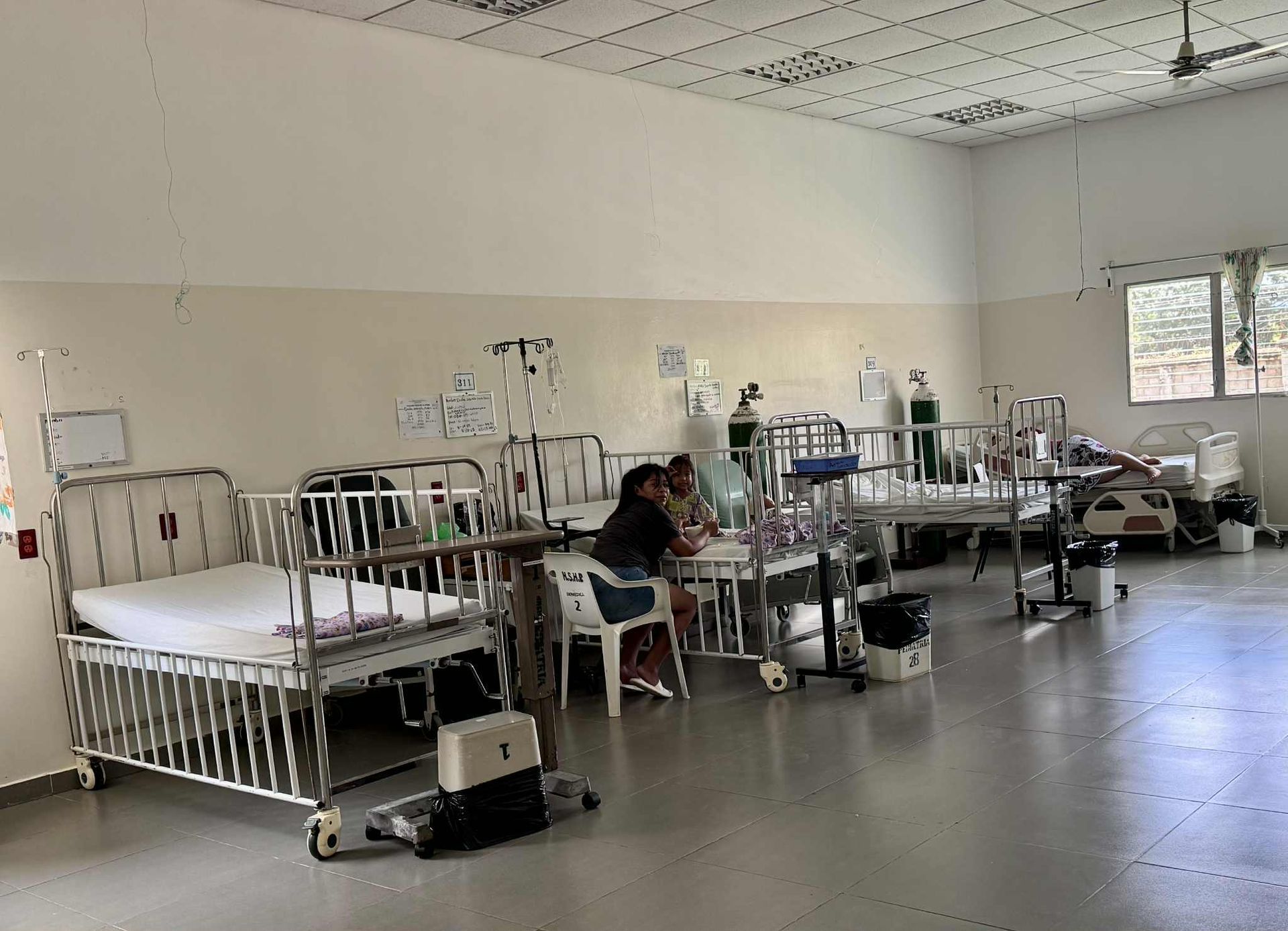Hospital room with several cribs. Two people sit near a crib. Oxygen tanks and medical equipment present.