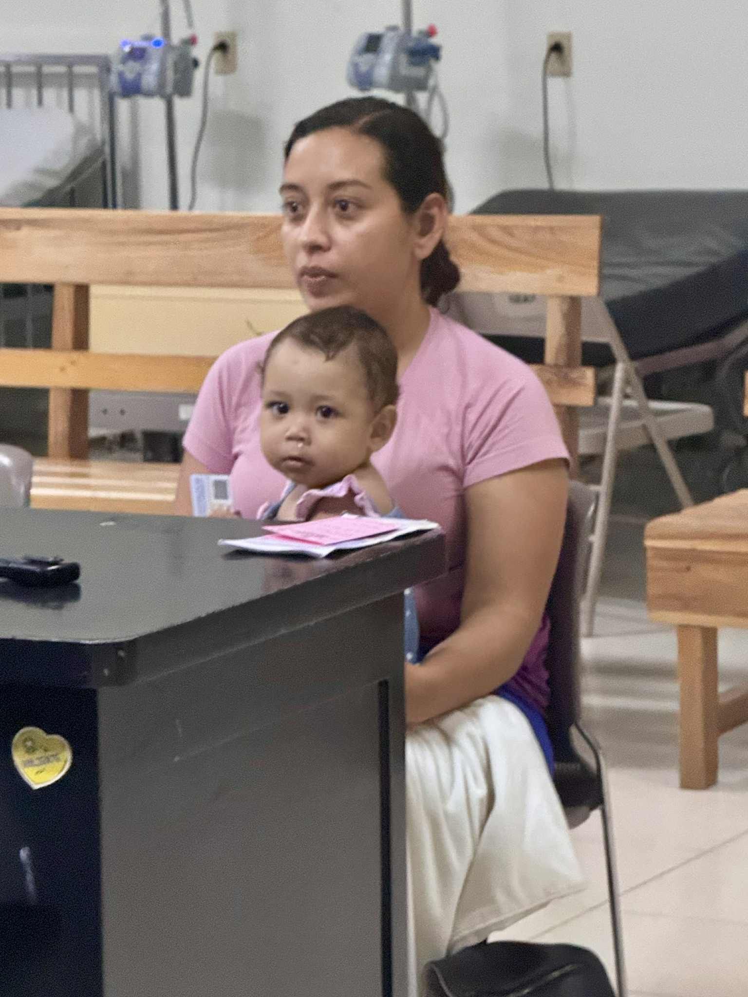 Woman holding a baby in a medical setting, both looking forward. The baby sits on a desk with papers.