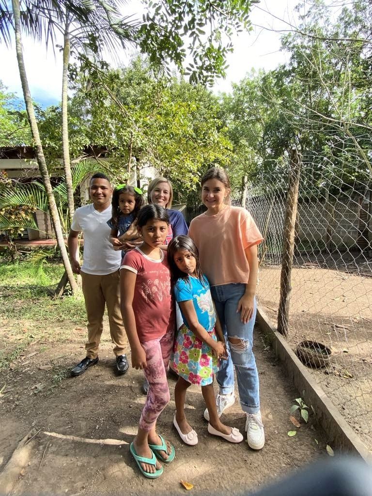 Group of people posing outdoors, including children and adults, near greenery.