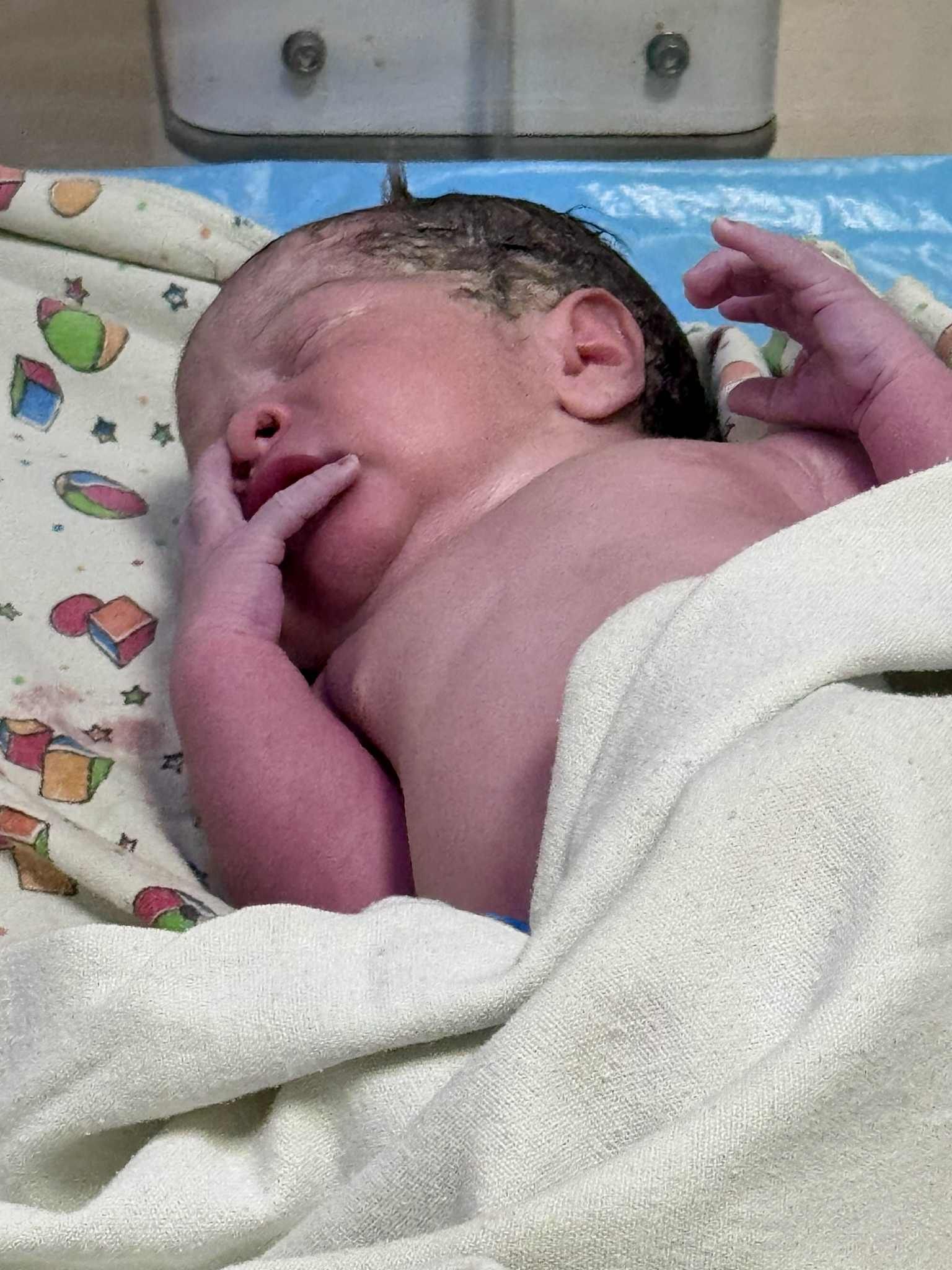 Newborn baby lying on a patterned sheet, wrapped in a towel, with eyes closed and hands near face.