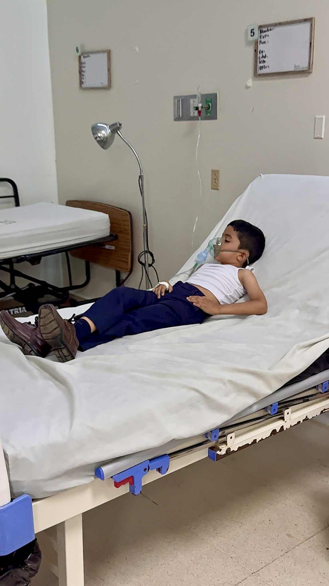 Boy lies on hospital bed, wearing face mask. Blue pants, white top. Medical setting.