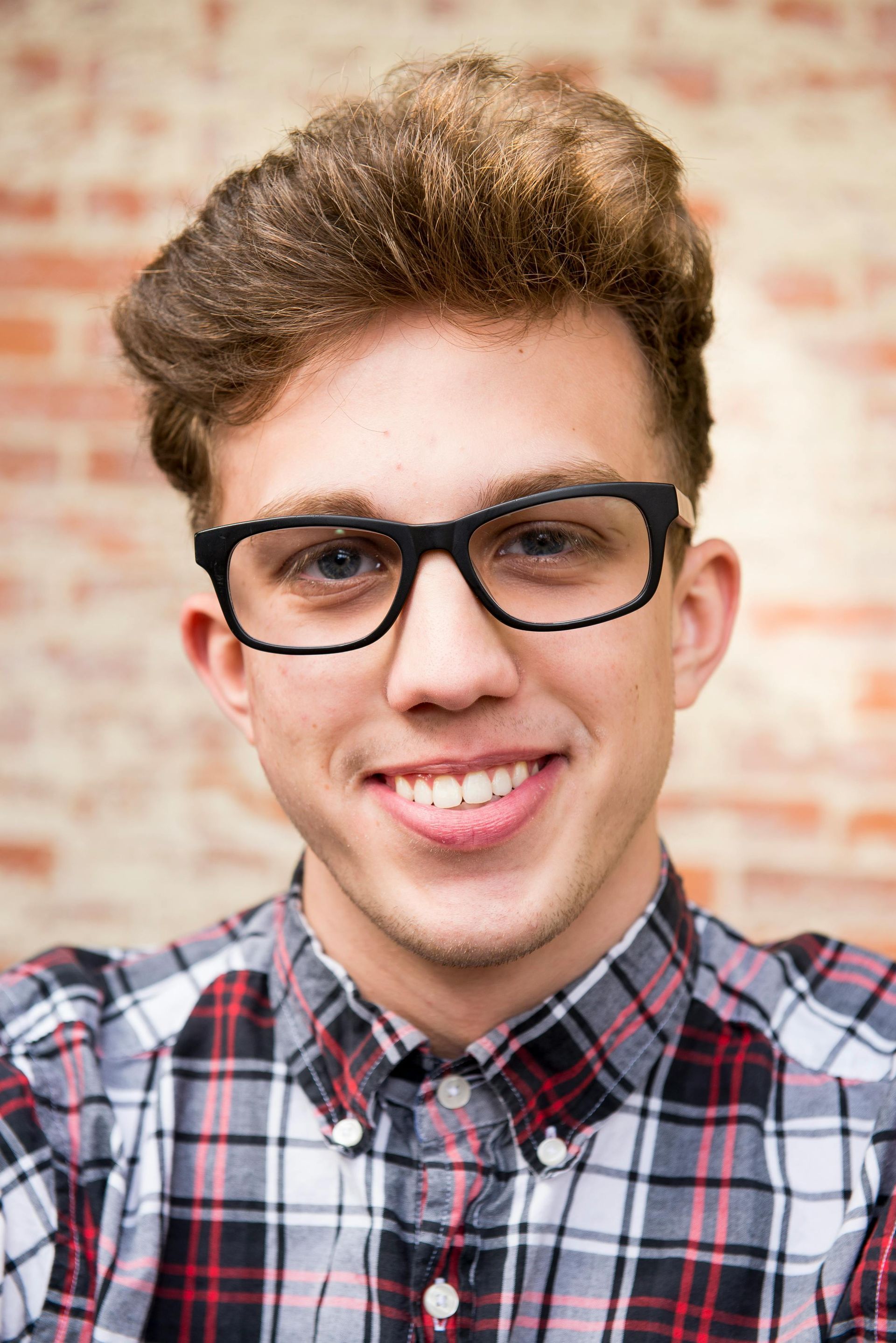 A young man wearing glasses and a plaid shirt is smiling.