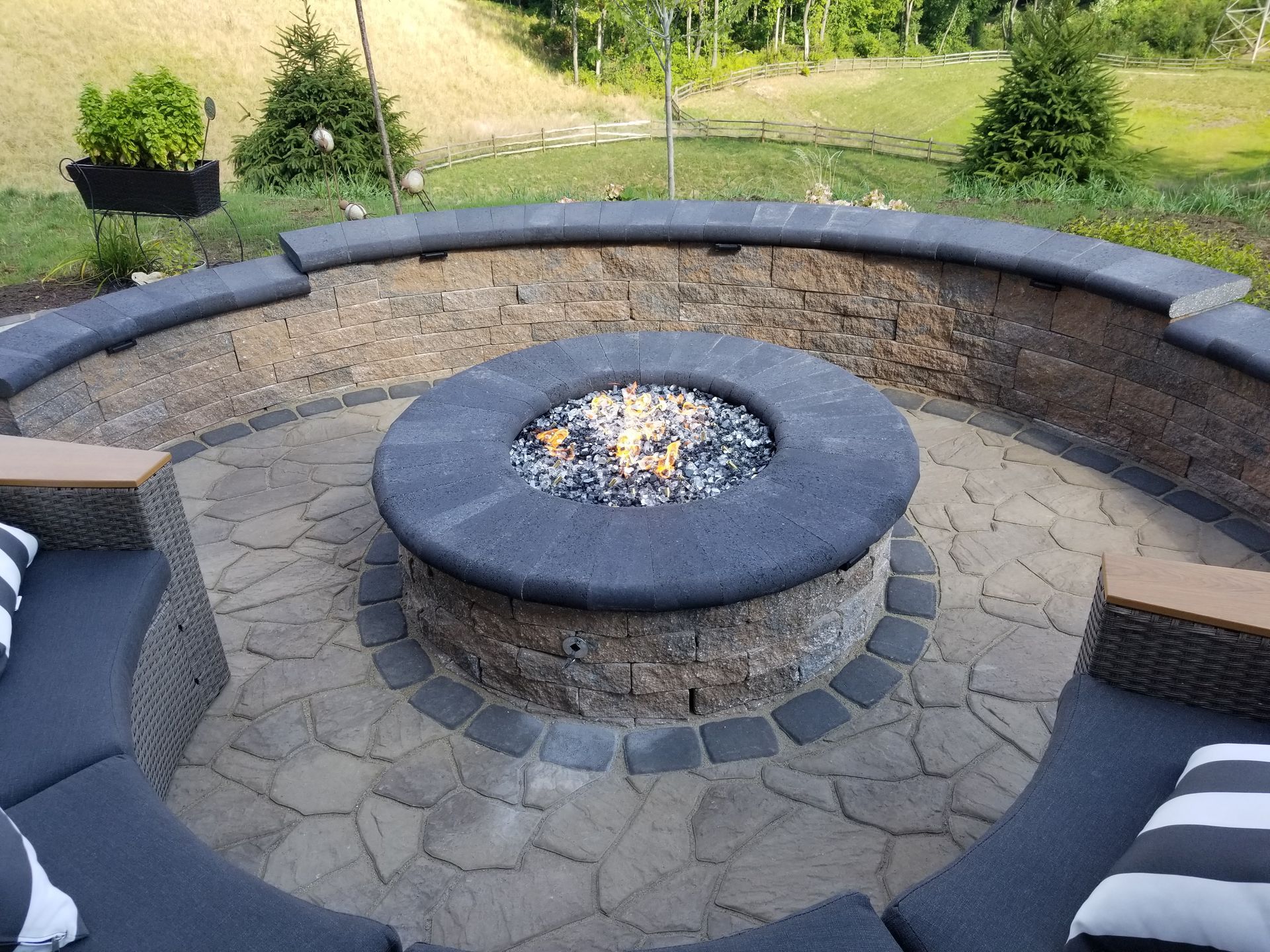 Outdoor stone fire pit with circular seating on a paved patio, surrounded by lawn and trees.