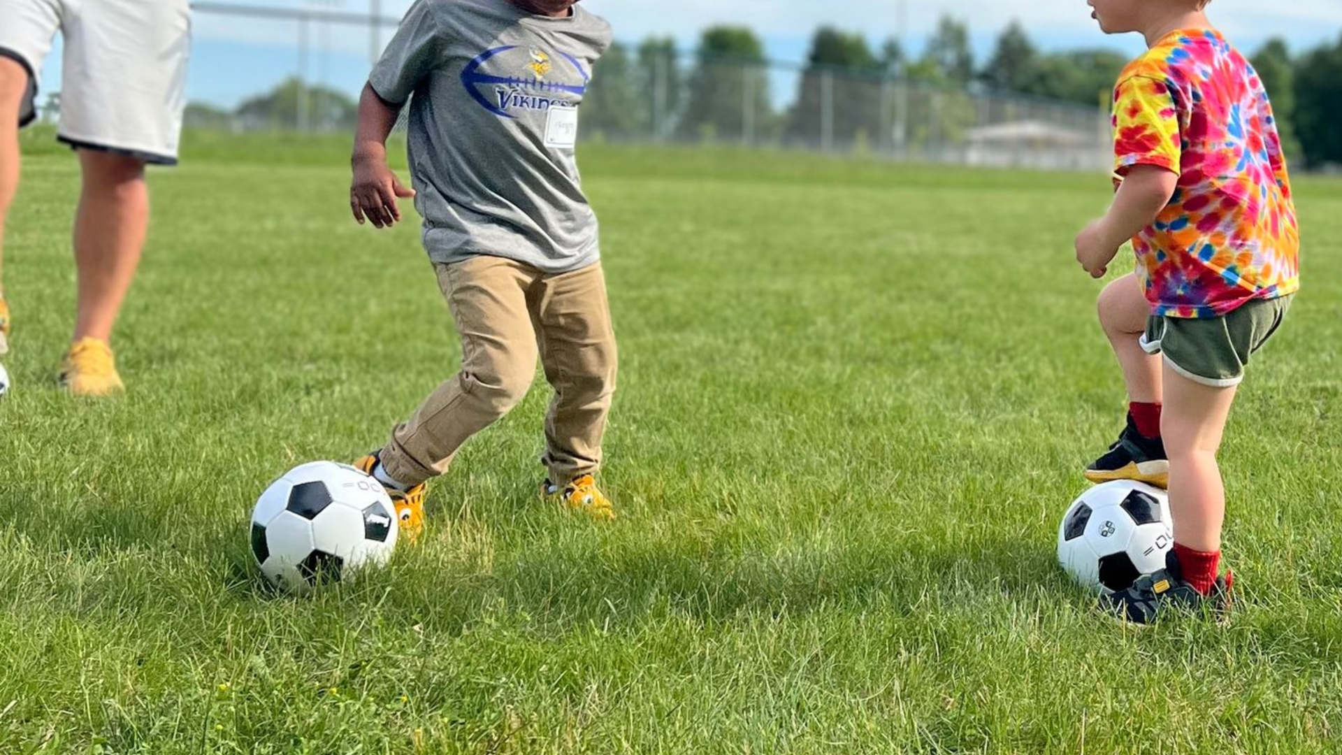 Two young children practicing soccer on a grass field with a coach guiding them during a Tiny Strikers MN session