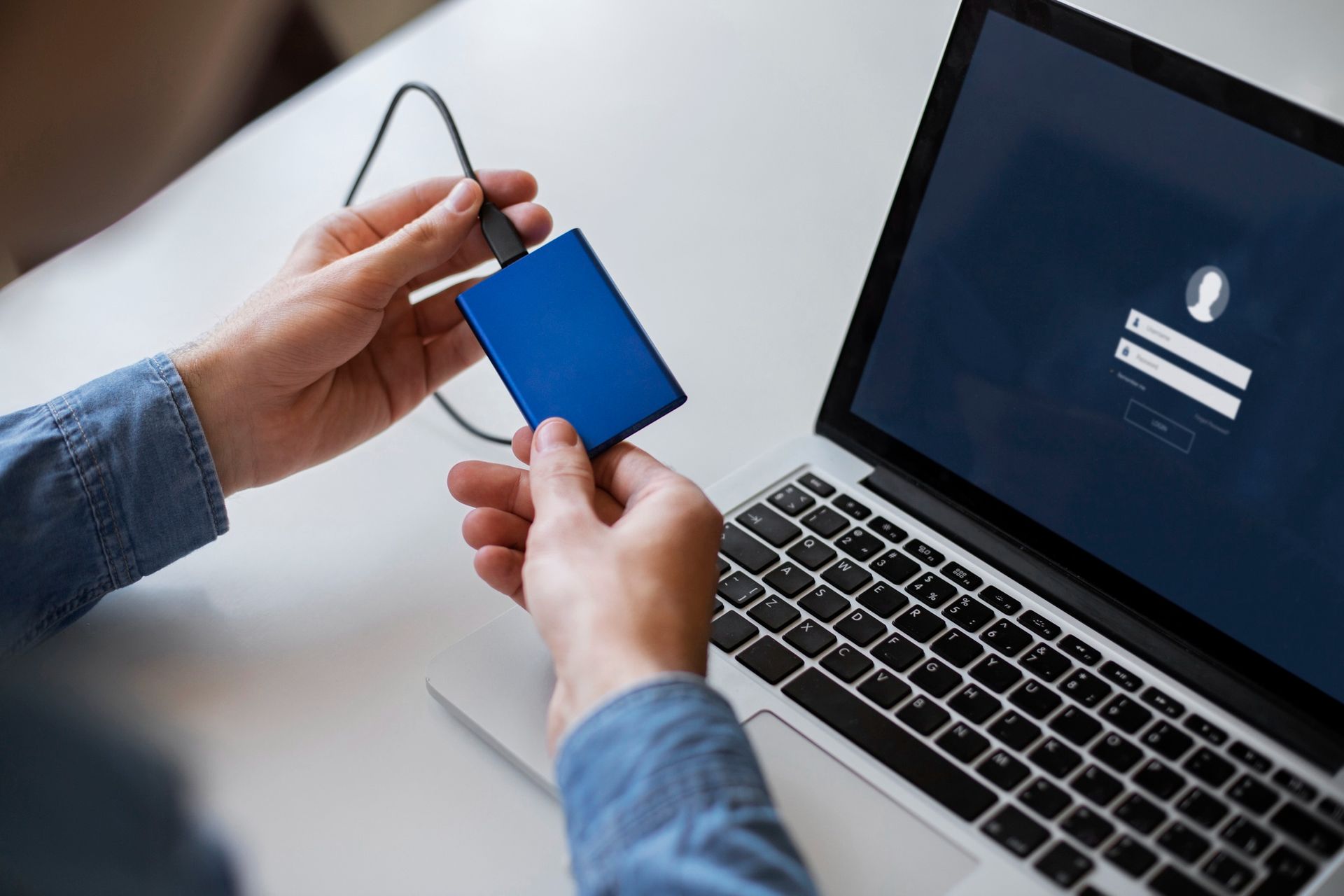 A person holds a blue external hard drive connected by a cable to a laptop displaying a login screen.