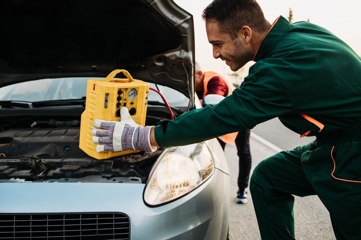 Man in green jumpsuit using a yellow jump starter to start a car with the hood open; another person in background.