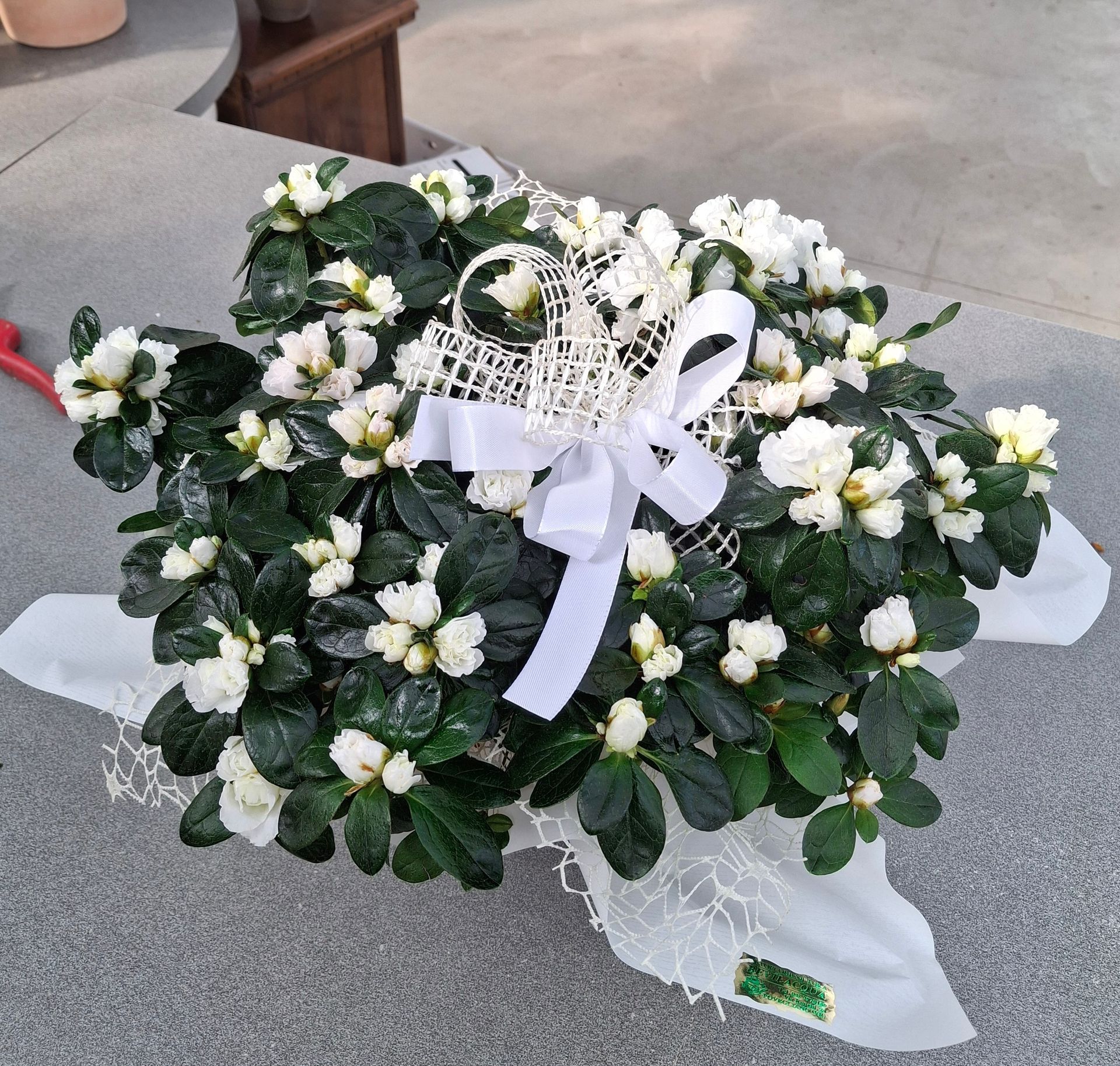 Composizione a forma di cuore di fiori bianchi con foglie verde scuro, legati con un nastro bianco e adagiati su un panno bianco.