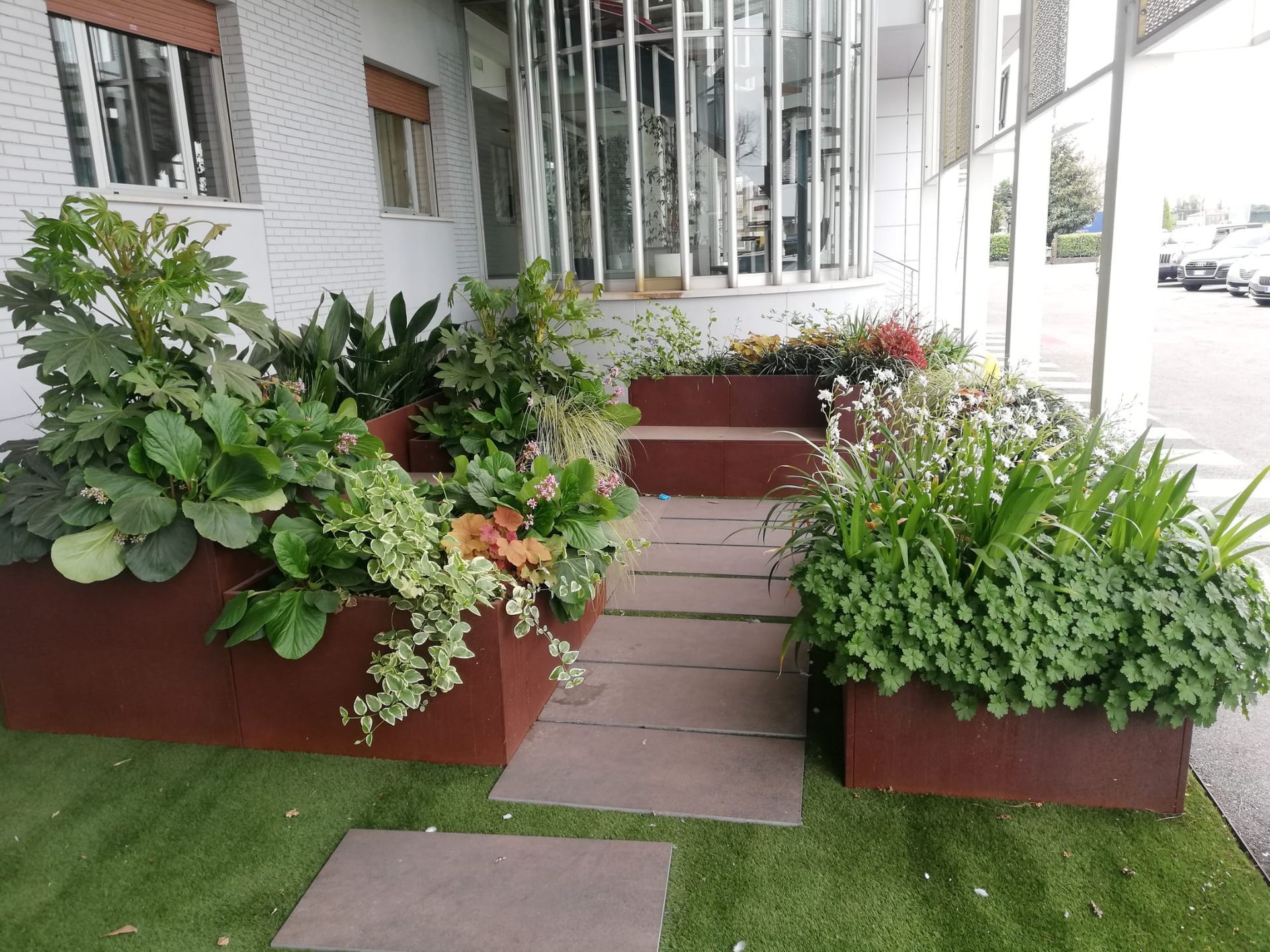 Ingresso esterno con fioriere rettangolari marroni piene di verde, che conduce a una porta a vetri sotto un edificio bianco.