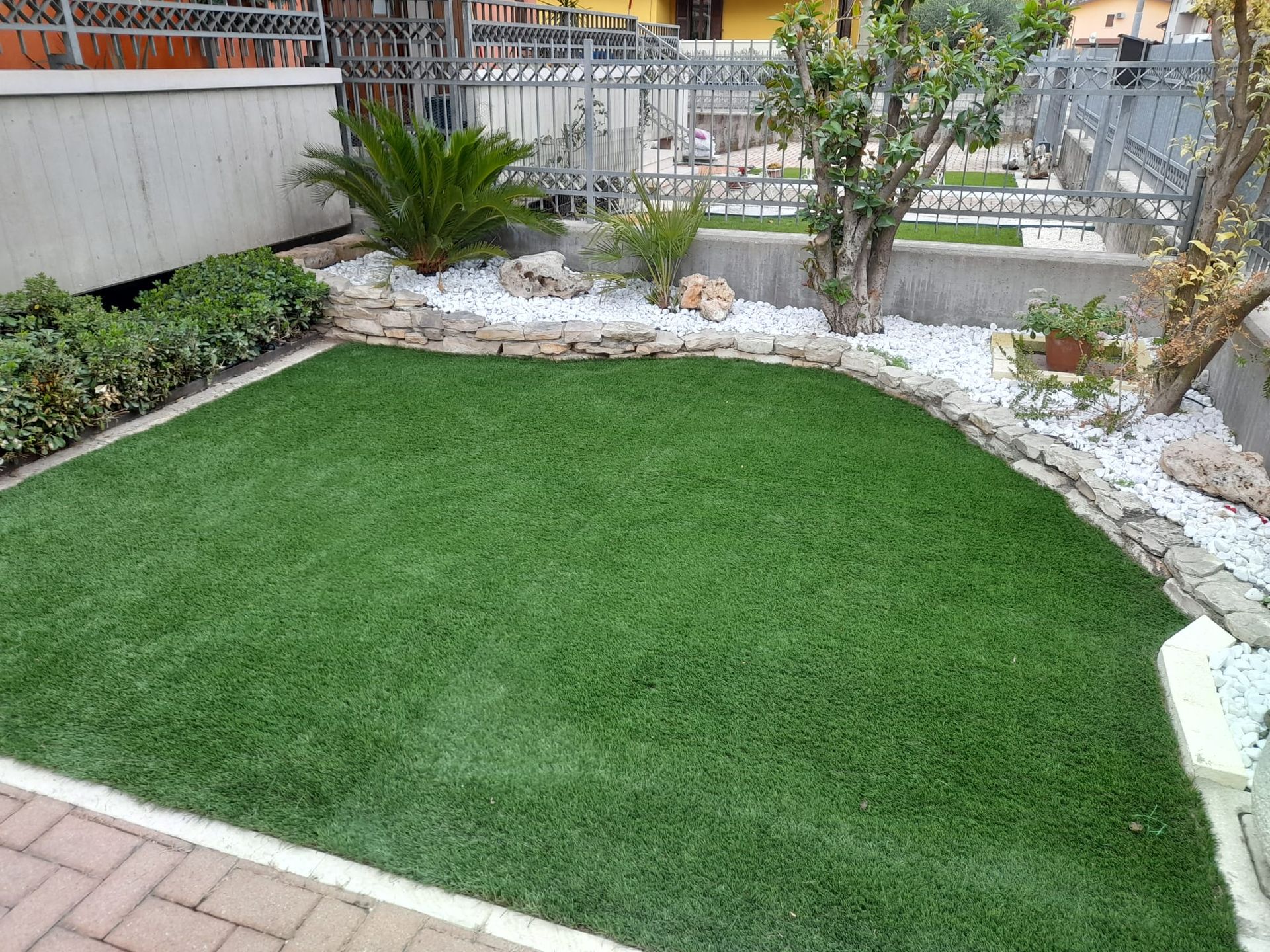 Un piccolo cortile ordinato con un appezzamento di erba sintetica verde circondato da piante, pietre bianche e una bordura di mattoni.