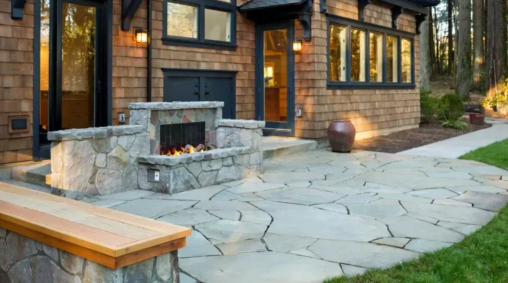 A stone patio with a fireplace and a bench in front of a brick house.