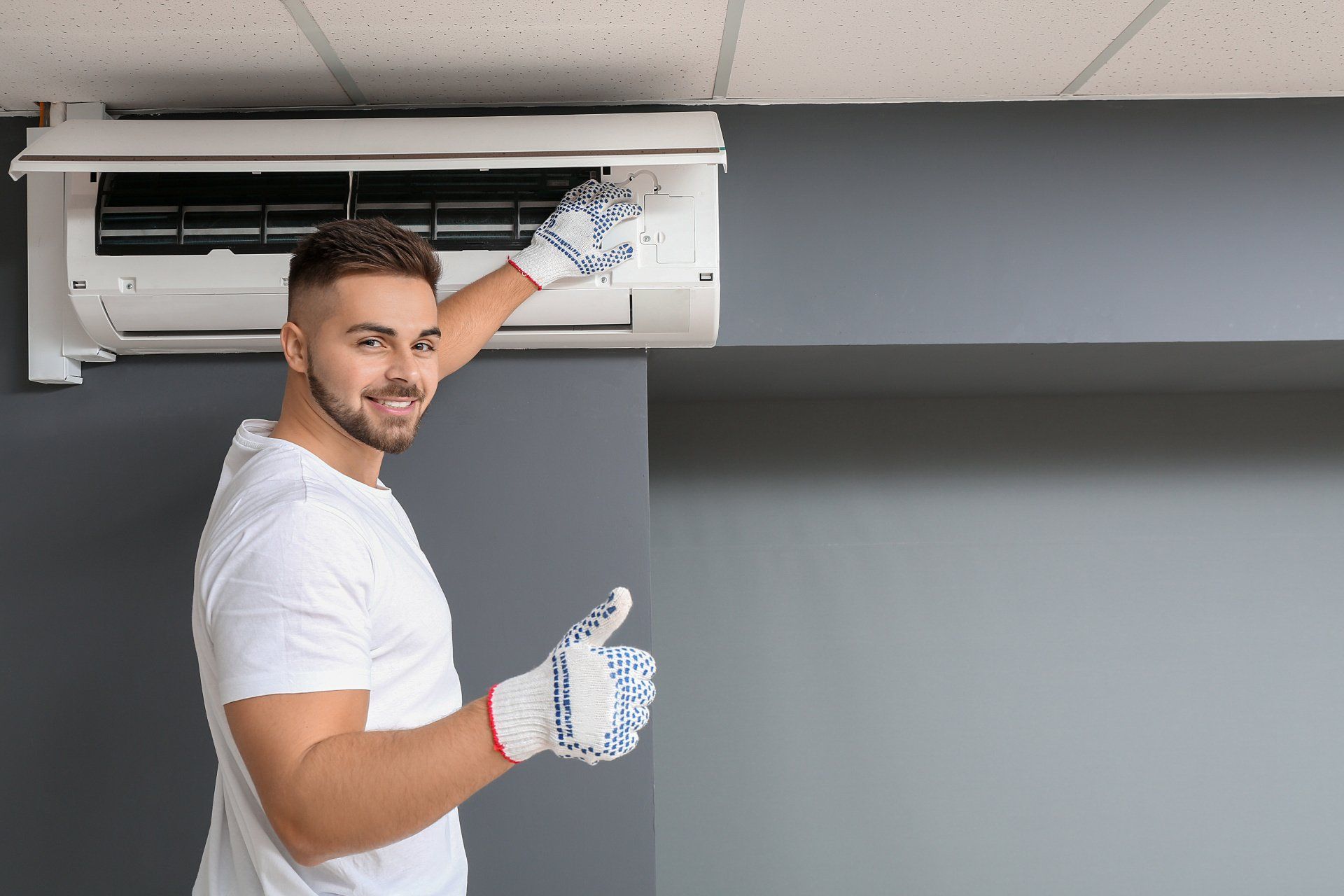 Man in gloves cleaning an AC unit with thumbs up in a room with gray walls.